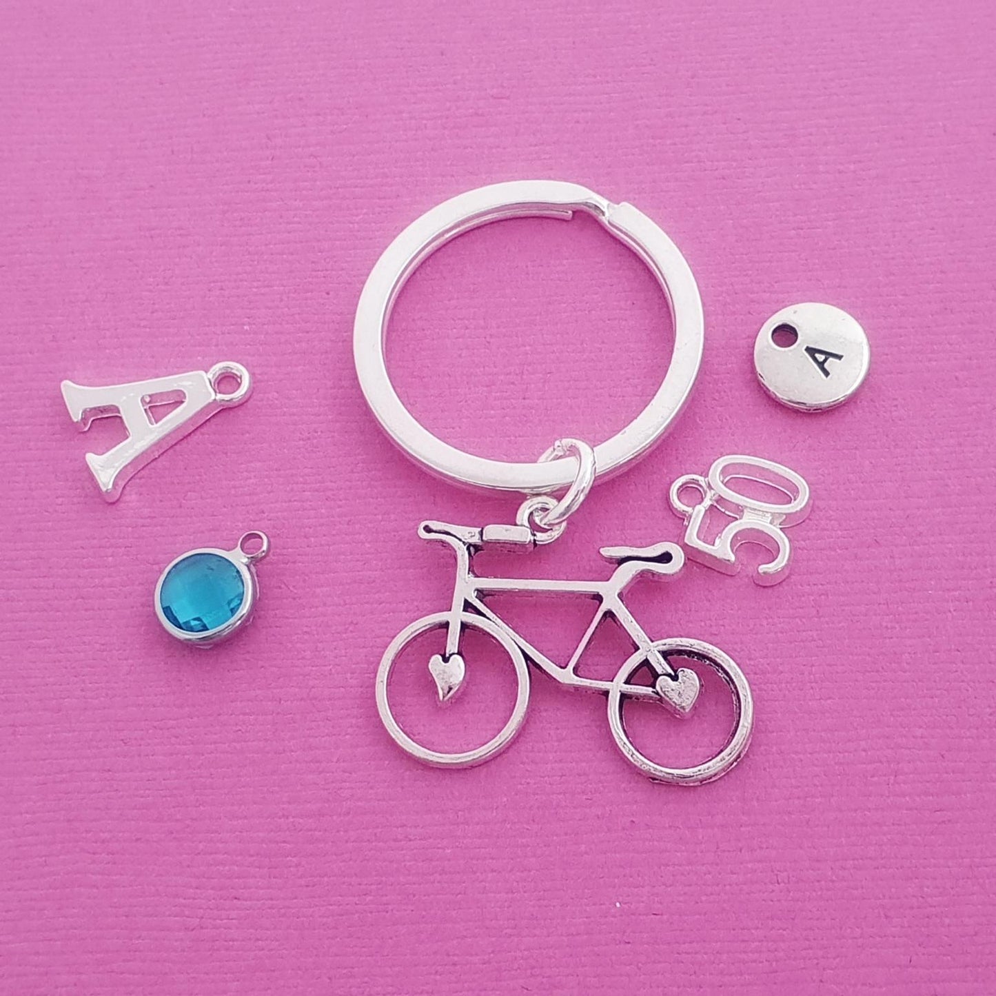 A silver keychain set featuring a bicycle charm, a split key ring, an initial 'A' charm, a small round charm with the letter 'A', and a blue circular charm, all arranged on a textured pink background.