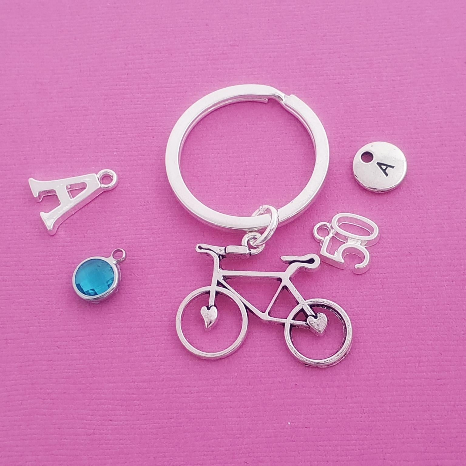 A silver keychain set featuring a bicycle charm, a split key ring, an initial 'A' charm, a small round charm with the letter 'A', and a blue circular charm, all arranged on a textured pink background.