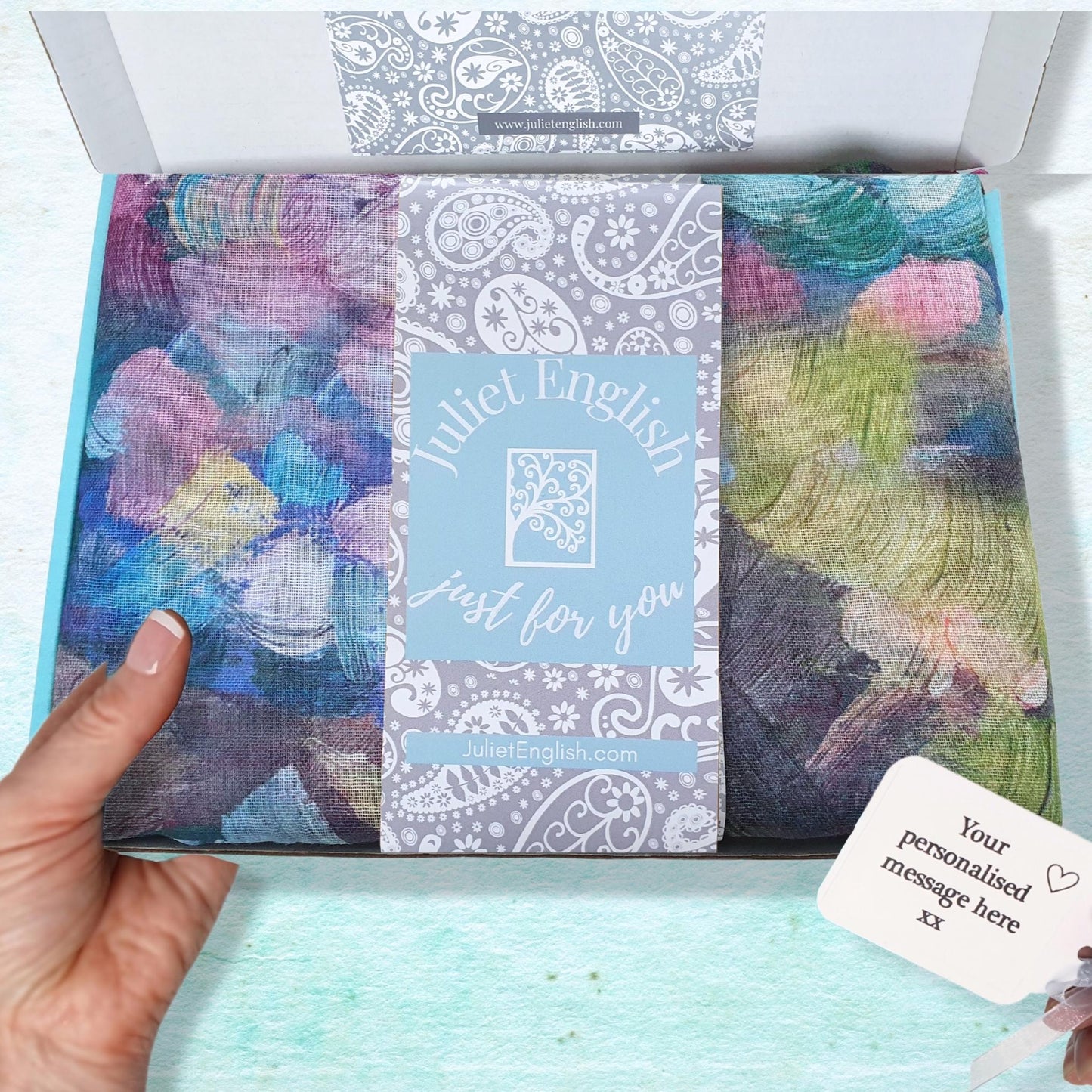 Scarf in a gift box with a visible brand label, held by a hand on a light blue background.