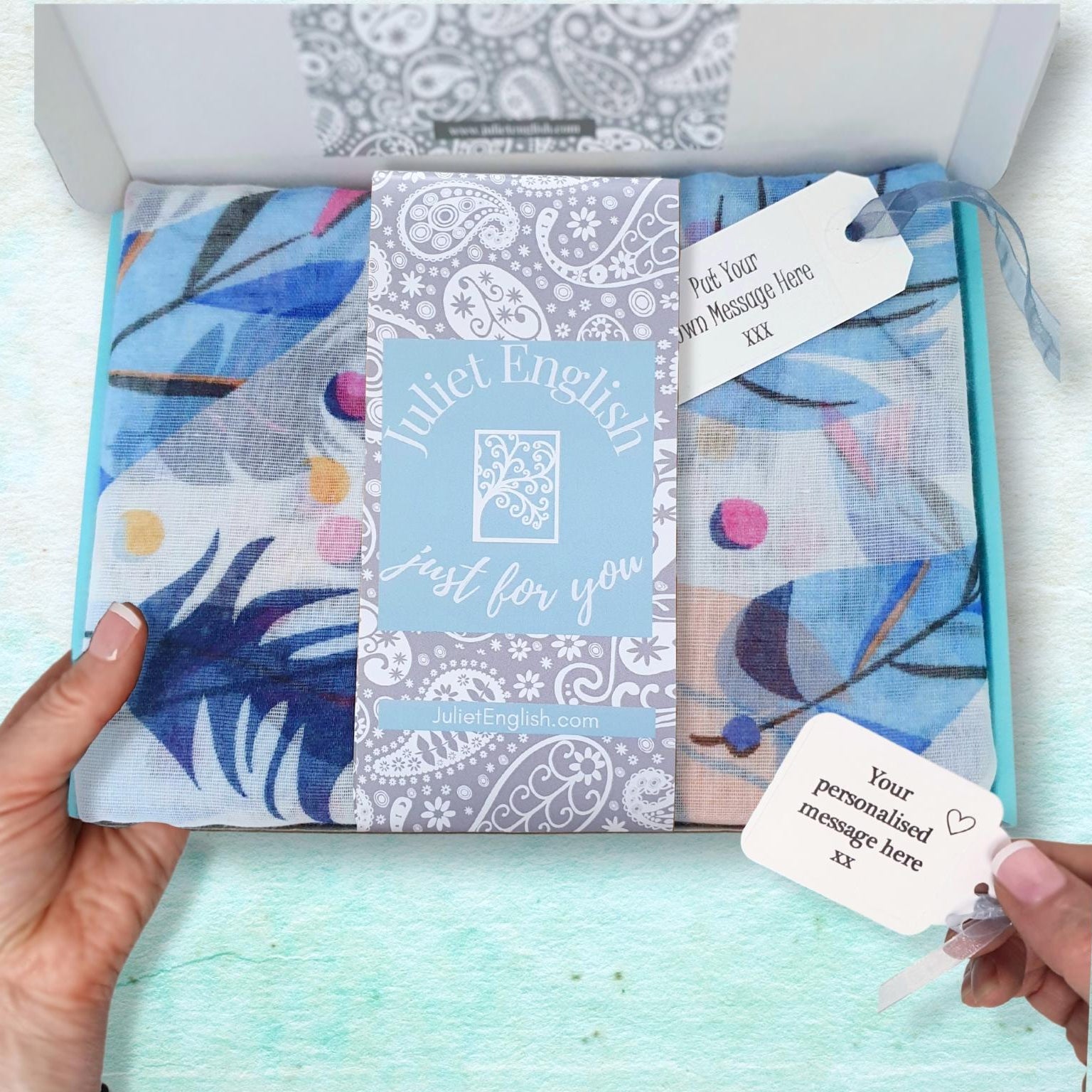 Packaged scarf with floral designs and personalised message tag, held by a hand against a light blue background.