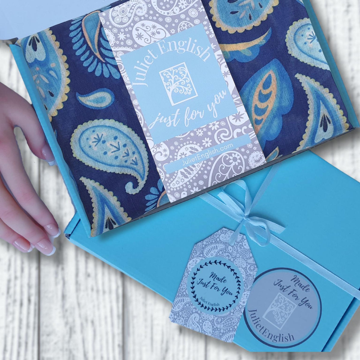 A turquoise blue gift box with a paisley patterned scarf and insert card and decorative tags reading 'Just for You', arranged on a white wood surface with a hand touching the box.