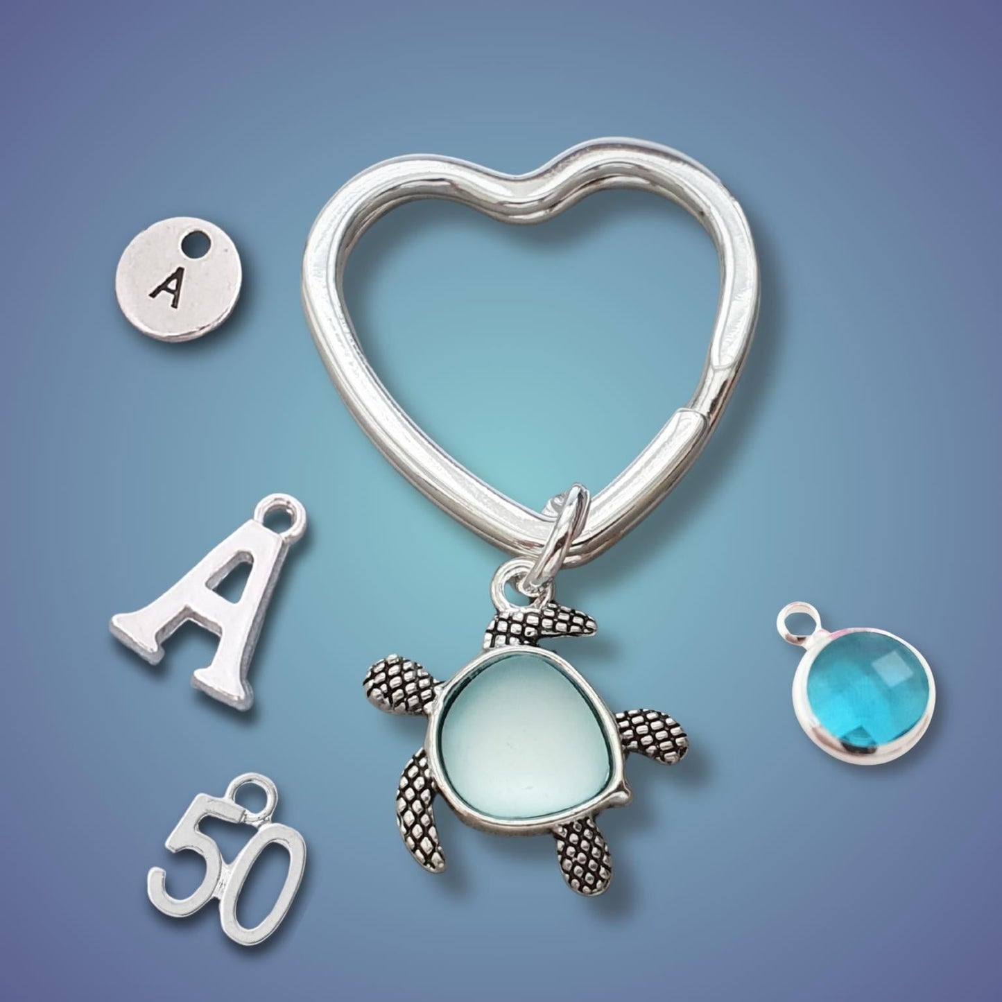 Silver heart-shaped key ring charm set with a sea turtle pendant, an initial 'A' disc charm, an initial 'A' letter charm, a '50' charm, and a round turquoise blue gemstone charm, displayed on a blue gradient background.