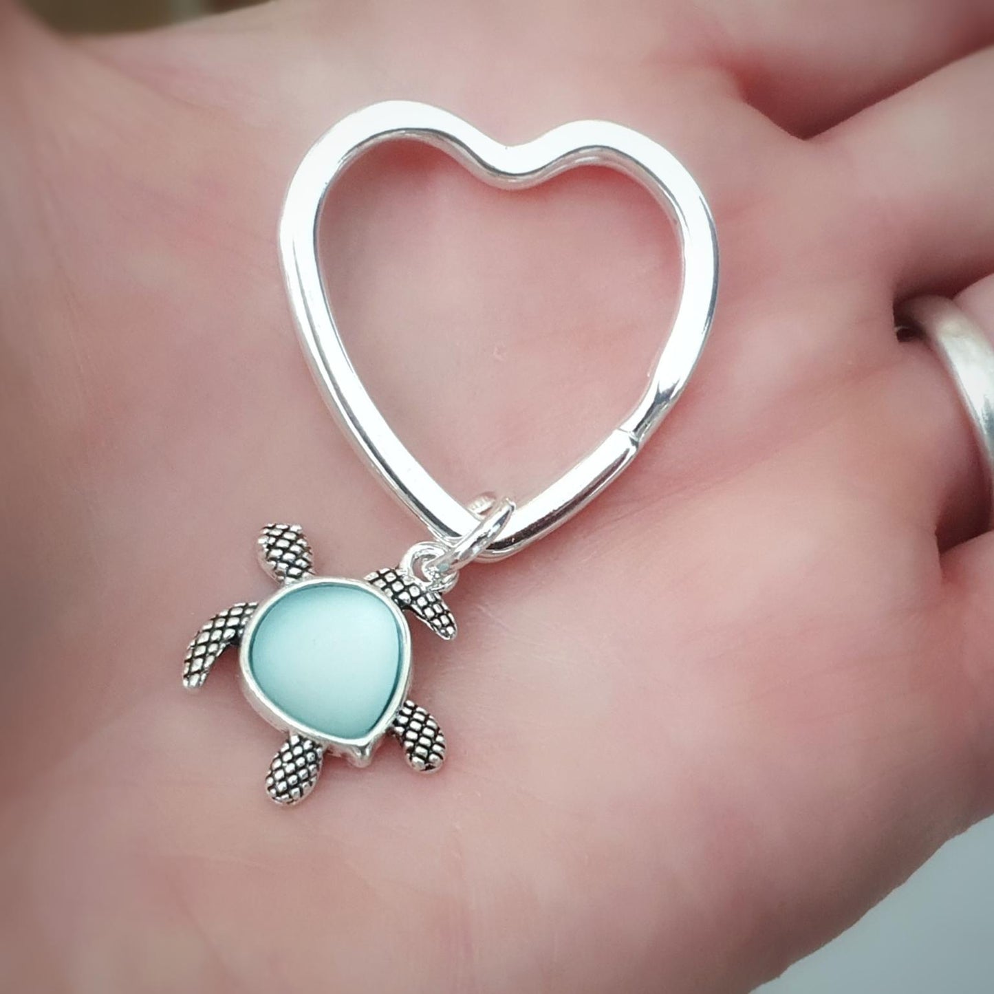 A silver heart-shaped key ring or bag charm with a small sea turtle pendant featuring a light blue oval center, displayed on a person's palm.