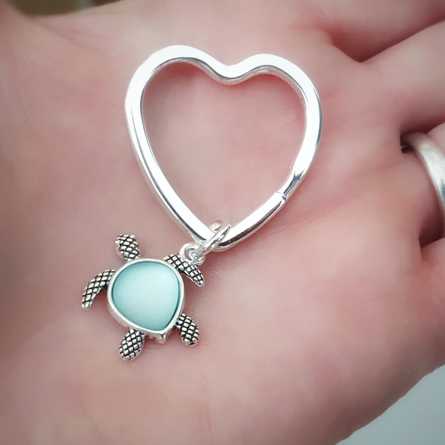 A silver heart-shaped key ring or bag charm with a small sea turtle pendant featuring a light blue oval center, displayed on a person's palm.