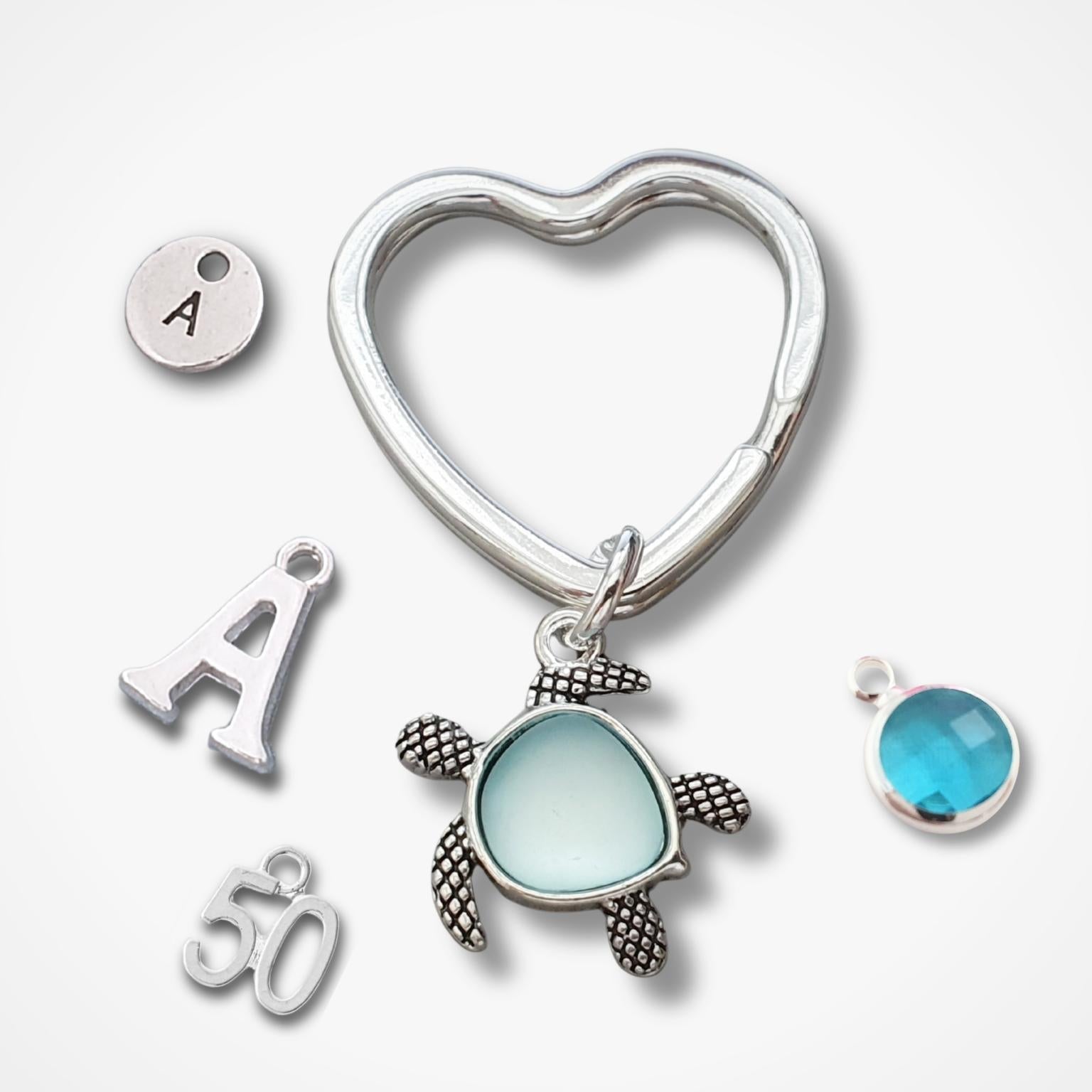 Silver jewelry charms arranged on a light gray background, including a heart-shaped keychain, a sea turtle charm with a light blue oval center, a round turquoise-blue bead charm, an initial 'A' letter charm, and a '50' charm.