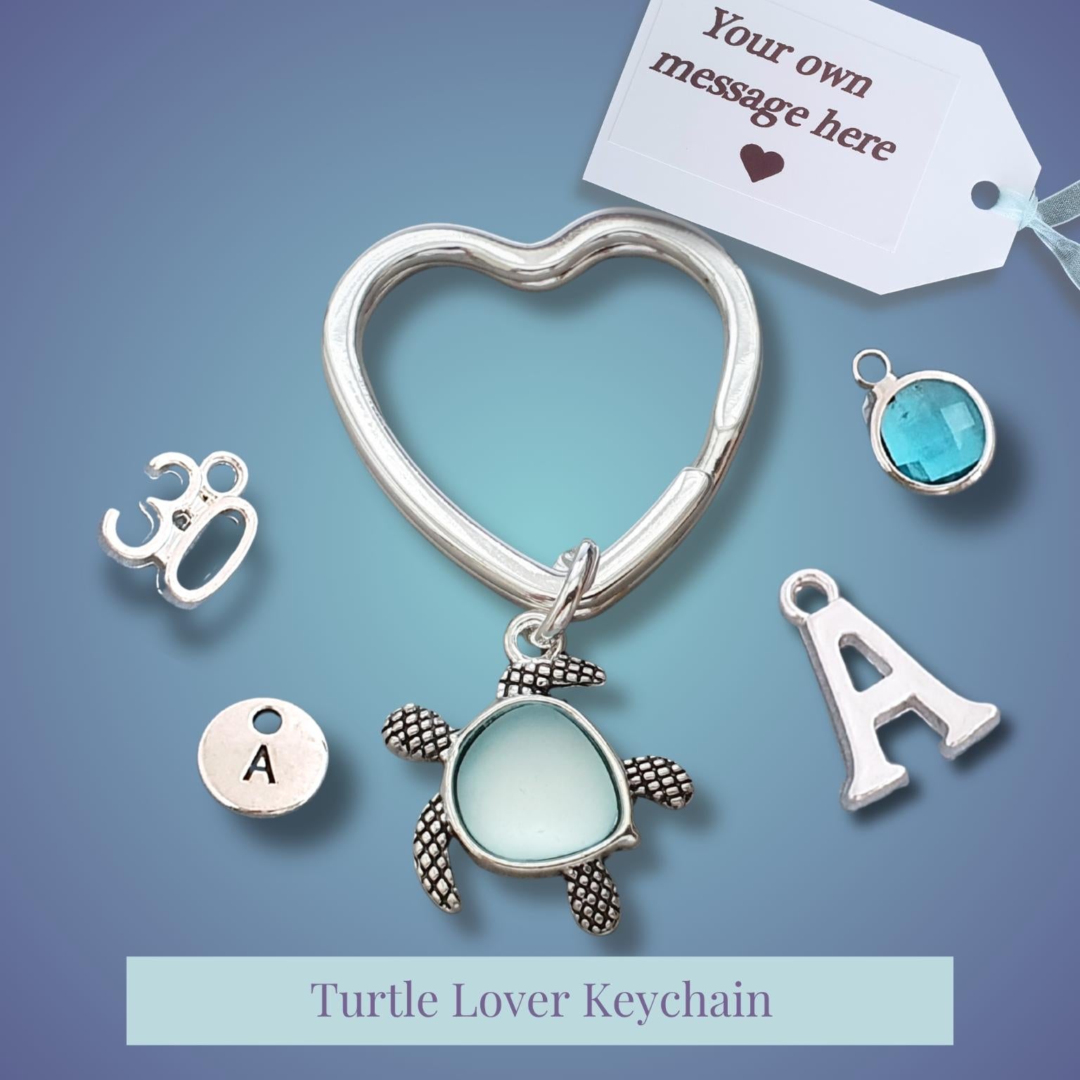 Silver heart-shaped keychain with a sea turtle charm and customizable letter and gemstone charms, displayed on a blue gradient background with a white tag that reads 'Your own message here'.
