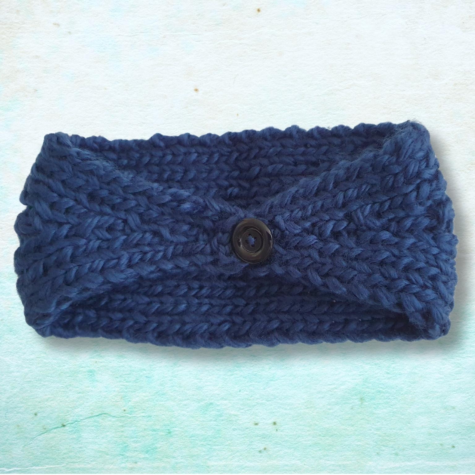 The back of a navy blue knitted headband with a central button closure, displayed flat against a textured light blue background.