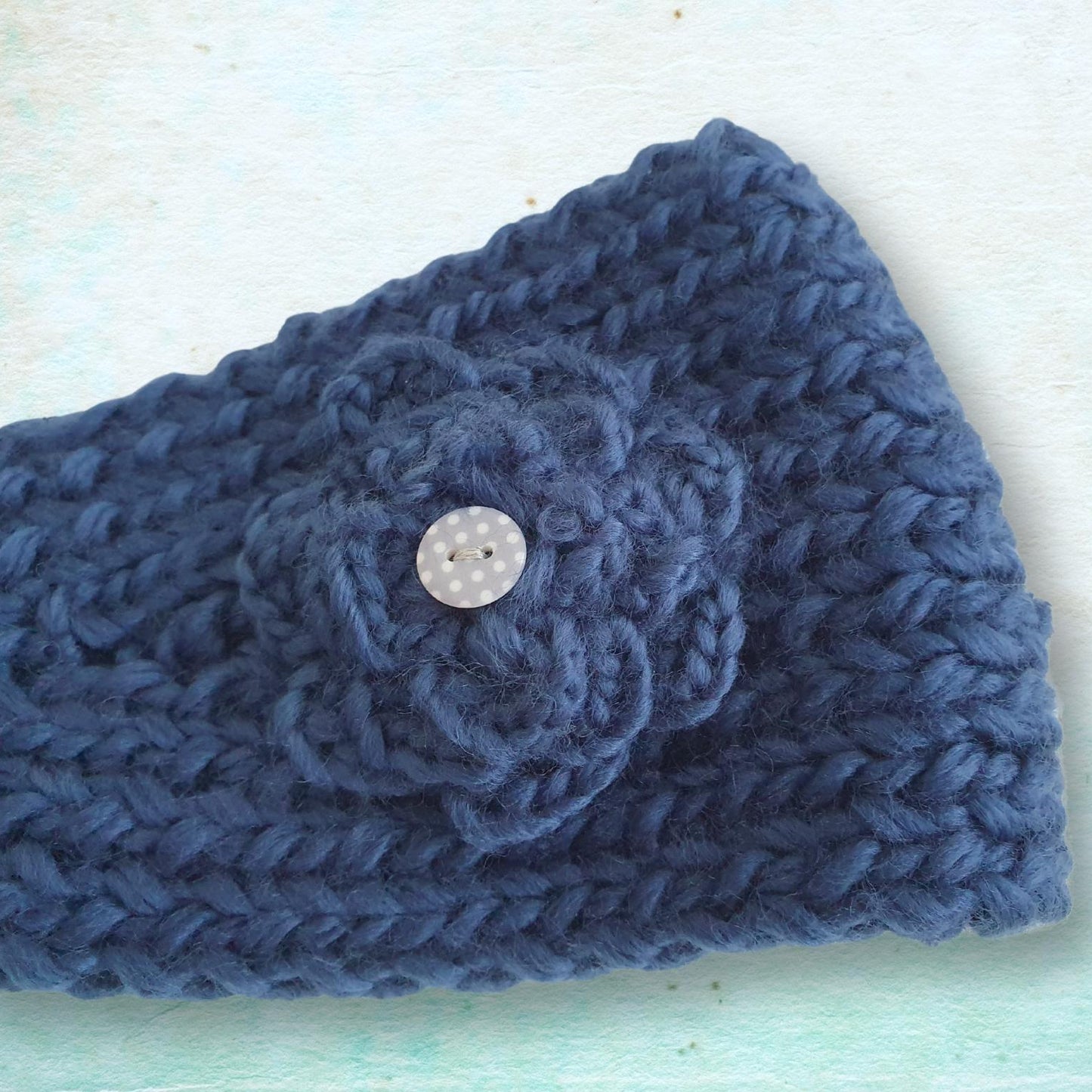 A close-up of a navy blue chunky knit headband featuring a crocheted flower applique and a small button, laid flat on a light textured background.