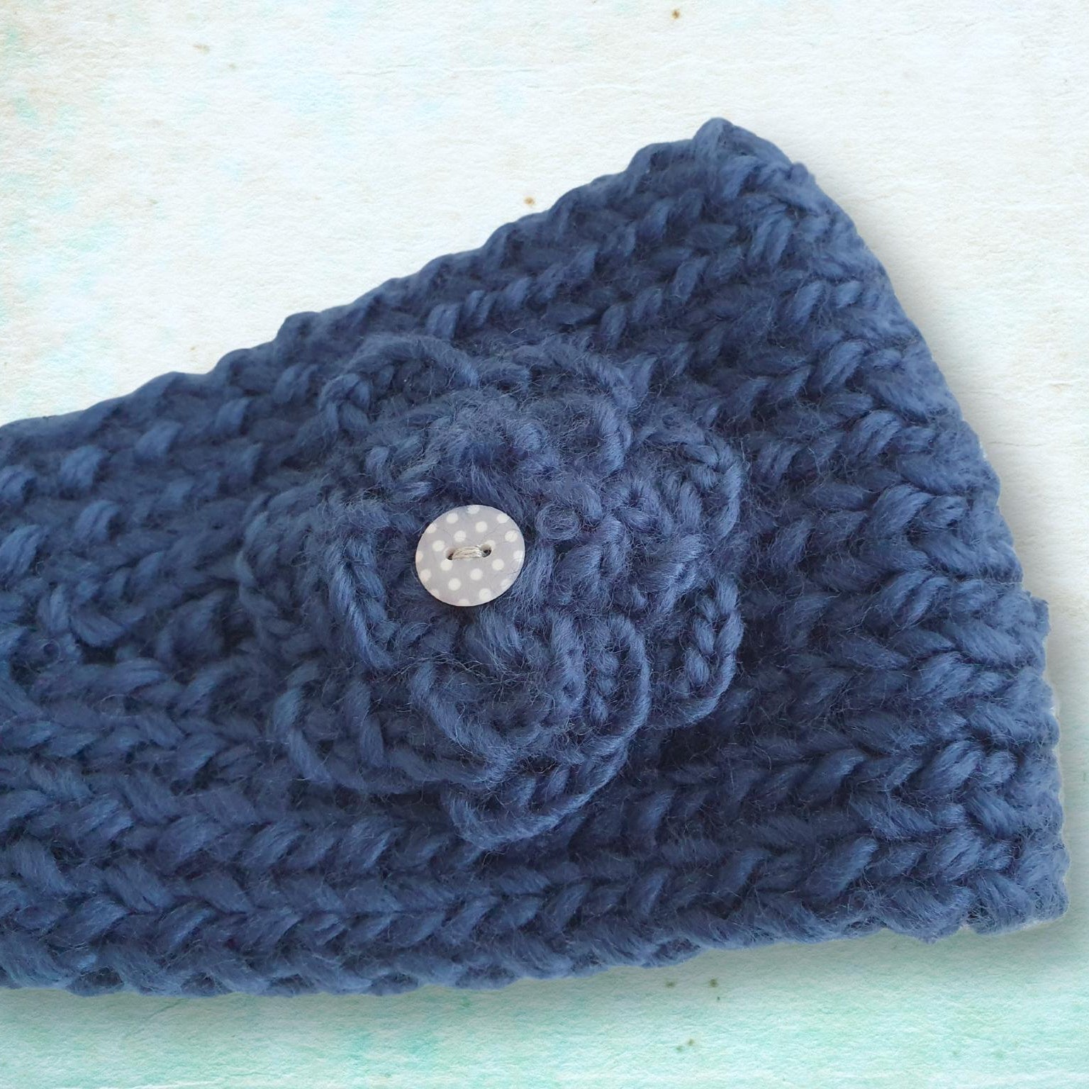A close-up of a navy blue chunky knit headband featuring a crocheted flower applique and a small button, laid flat on a light textured background.