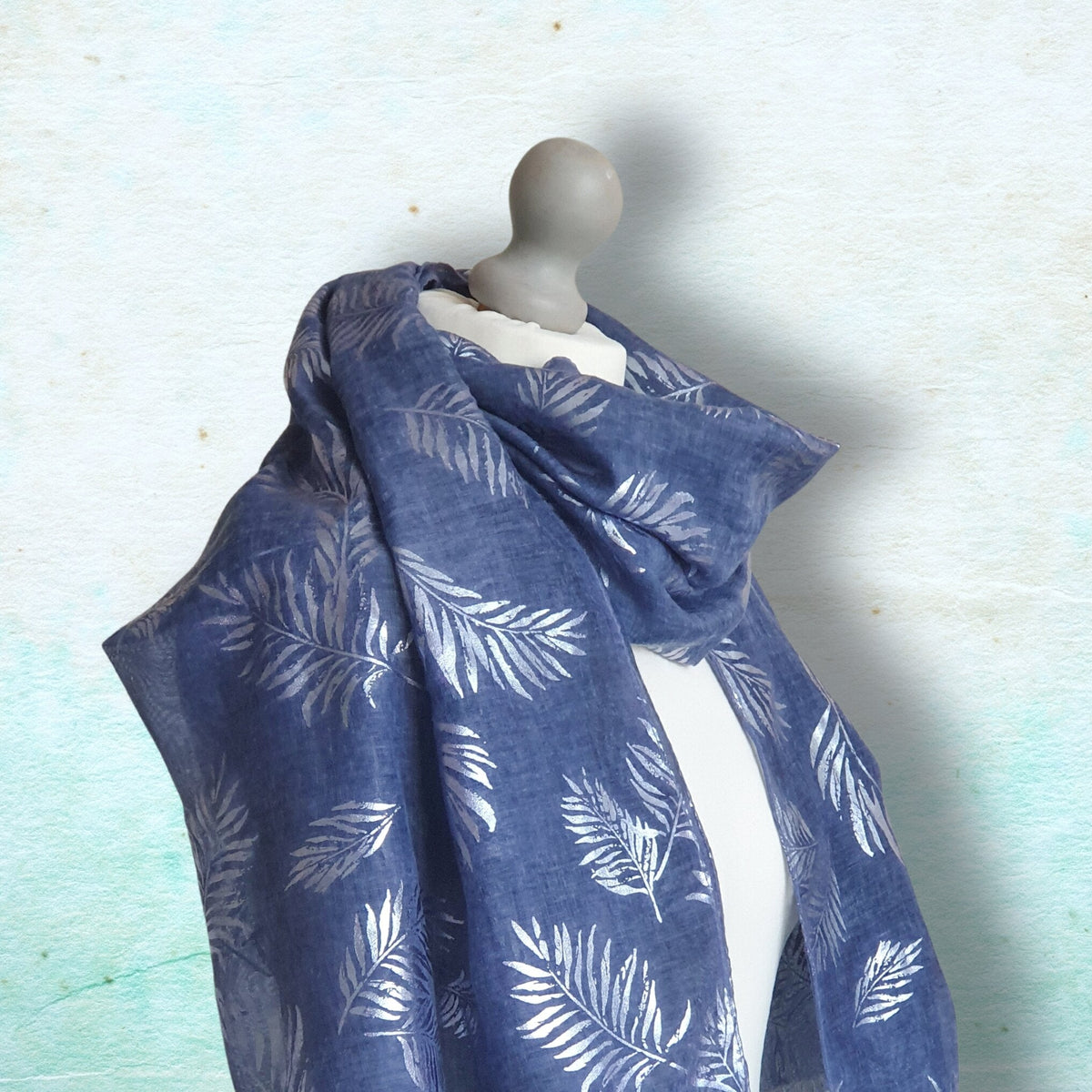 A blue scarf with a white palm leaf pattern draped on a gray mannequin torso against a textured light blue background.