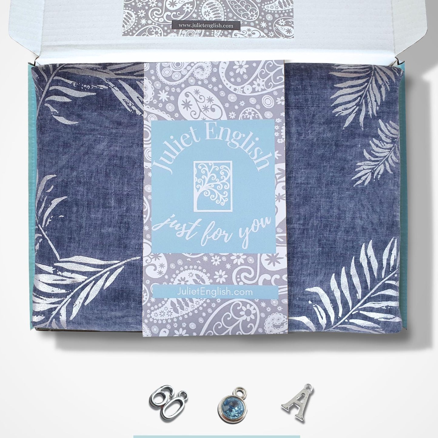 An open gift box from Juliet English on a white background, containing a blue and white patterned fabric scarf with the brand name and a small silver charm set below.