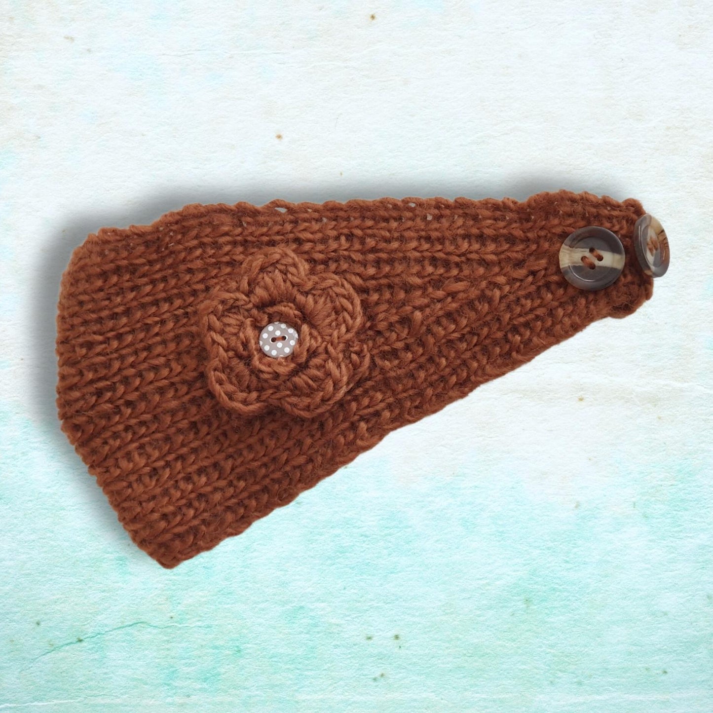 A brown knitted headband with a crocheted flower applique and two wooden buttons, displayed on a textured light blue background.