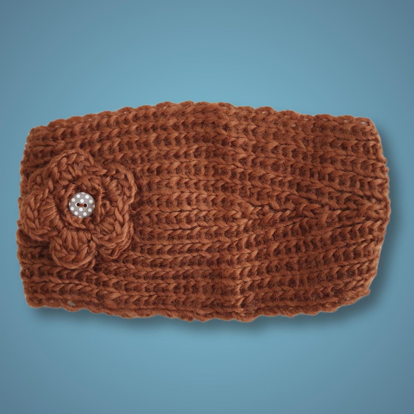 A brown chunky knit headband featuring a textured flower applique on the side with a button-embellished center, displayed against a solid blue background.