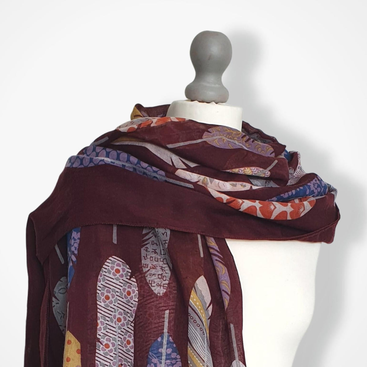 A multicolor scarf with a deep burgundy base and feather patterns, draped on a white mannequin torso against a white background.