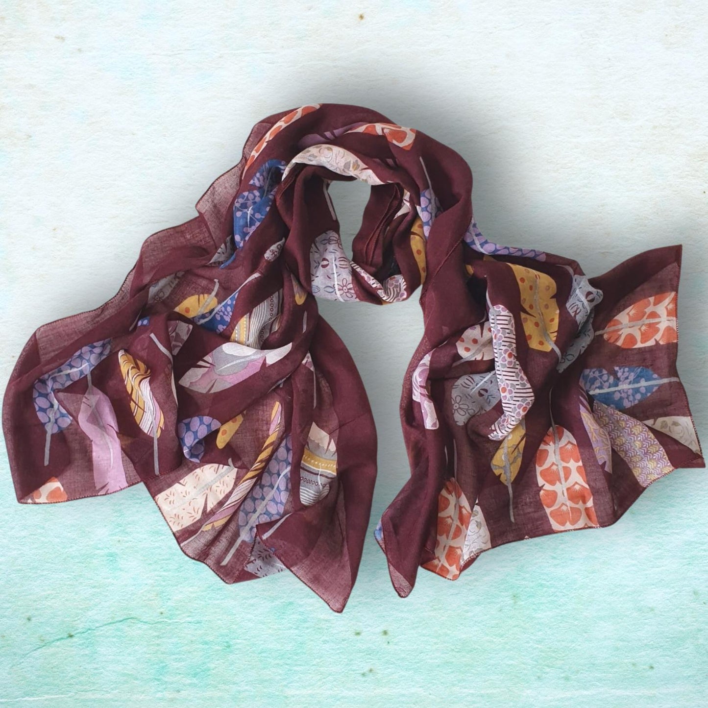 A lightweight, semi-sheer scarf with a multicolor pattern of feathers and abstract shapes in shades of burgundy, pink, orange, yellow, and blue, laid flat on a textured light blue-green background.