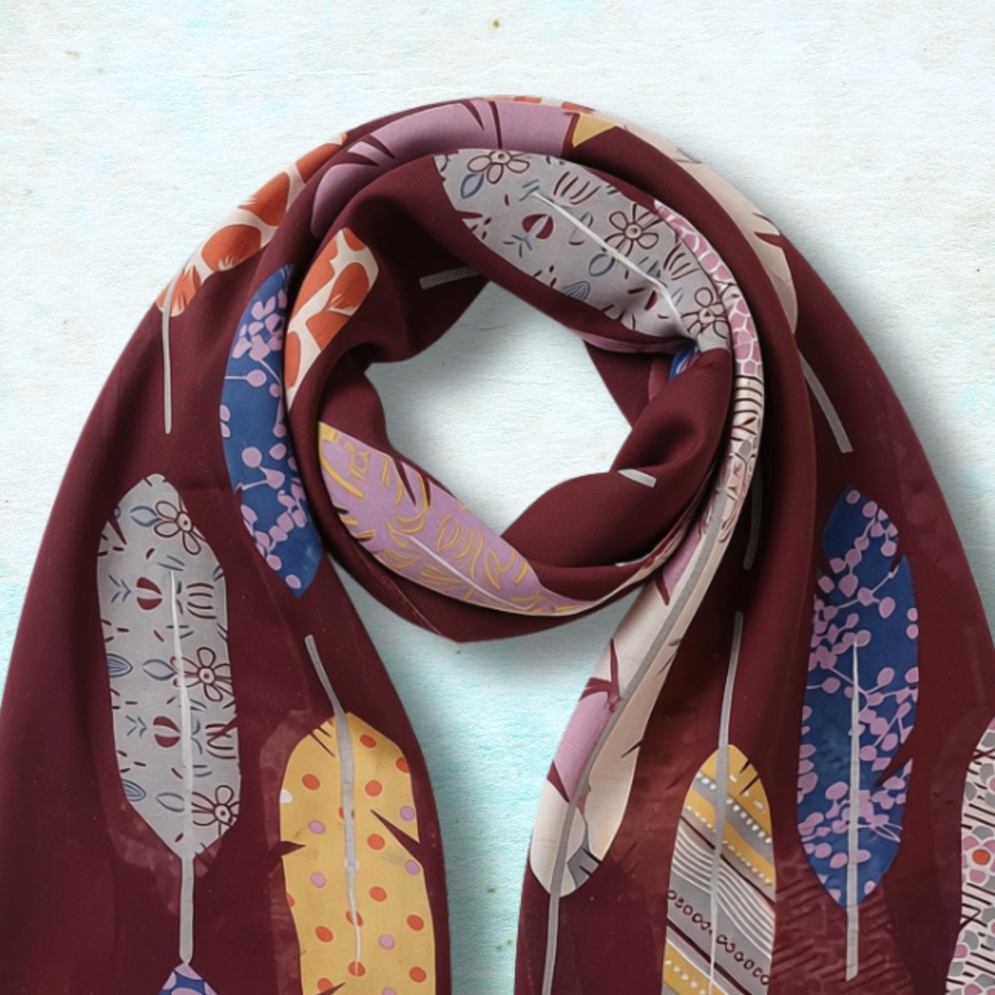 A long scarf with a deep burgundy base and a colorful feather pattern in yellow, blue, pink, and white, styled in a loop on a textured light blue background.