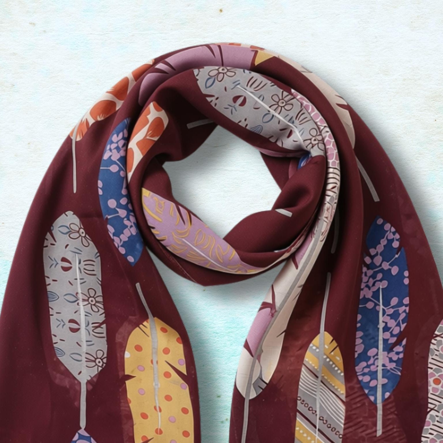 A long scarf with a deep burgundy base and a colorful feather pattern in yellow, blue, pink, and white, styled in a loop on a textured light blue background.