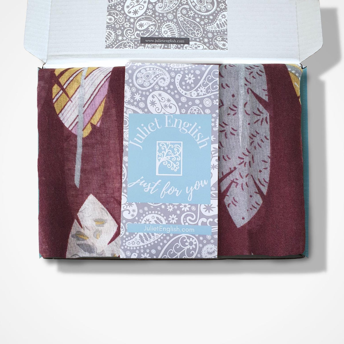 A boxed scarf displayed inside an open white box on a light grey background. The scarf is burgundy, lavender, white, and yellow feather-like prints, with a central light blue card reading 'Juliet English just for you.