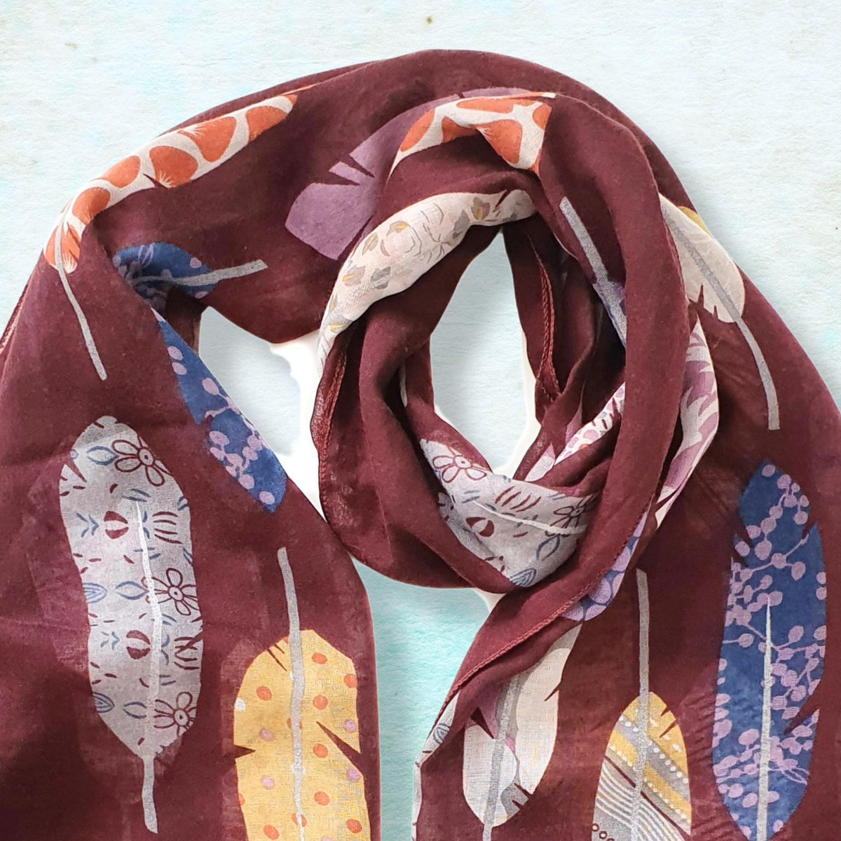 A lightweight sheer scarf in deep burgundy, featuring a colorful print of stylized feathers in blue, yellow, orange, and white, draped on a light blue textured background.