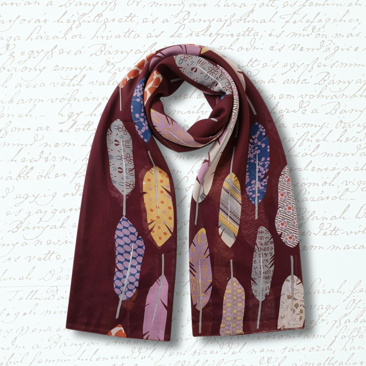 A long scarf in deep burgundy with a pattern of colorful feathers in pink, yellow, blue, and gray, displayed against a light background with faint cursive writing.