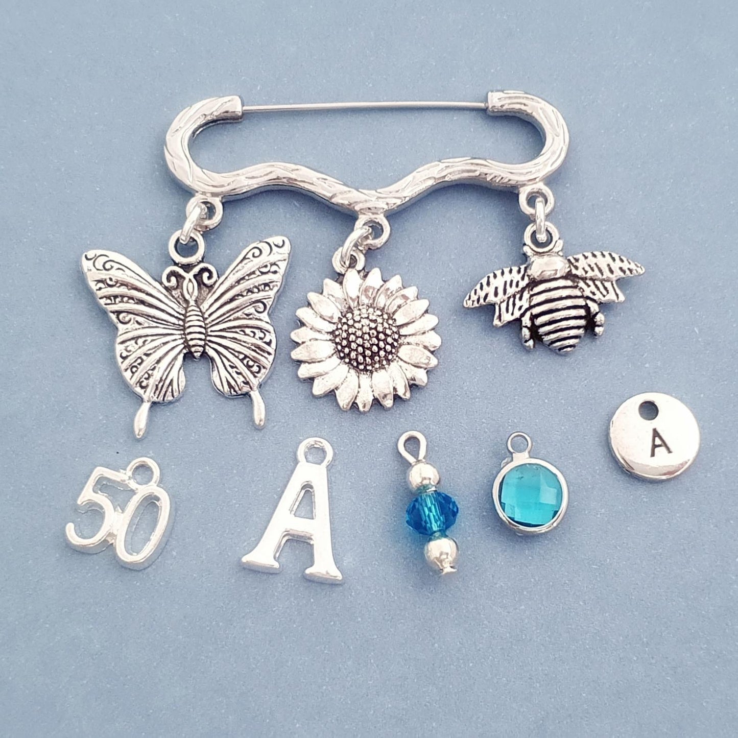 Brooch with silver charms including a butterfly, sunflower, bee, number 50, letter 'A', and blue beads on a light blue background.