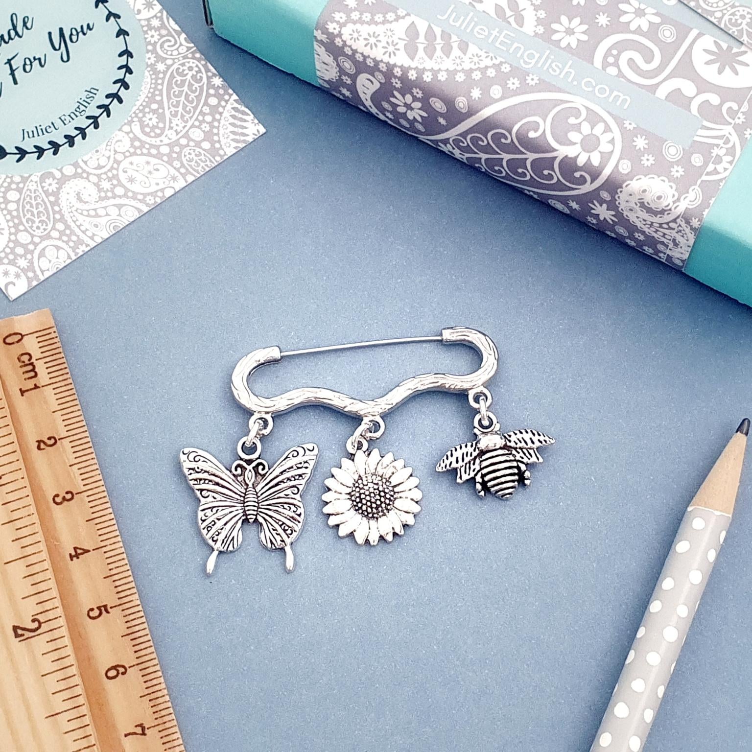 Silver brooch with butterfly, sunflower, and bee charms on a blue surface with a ruler and box.