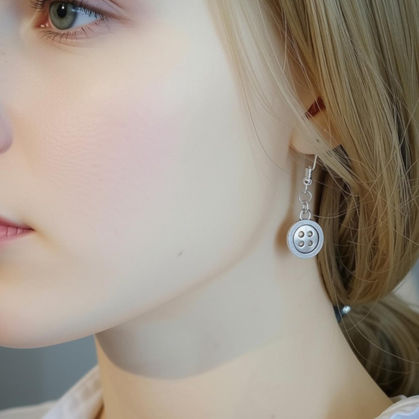 A close-up of a woman's ear wearing a silver button dangle earring. The earring features a round silver button charm with four buttonholes, suspended from a delicate silver wire hook.