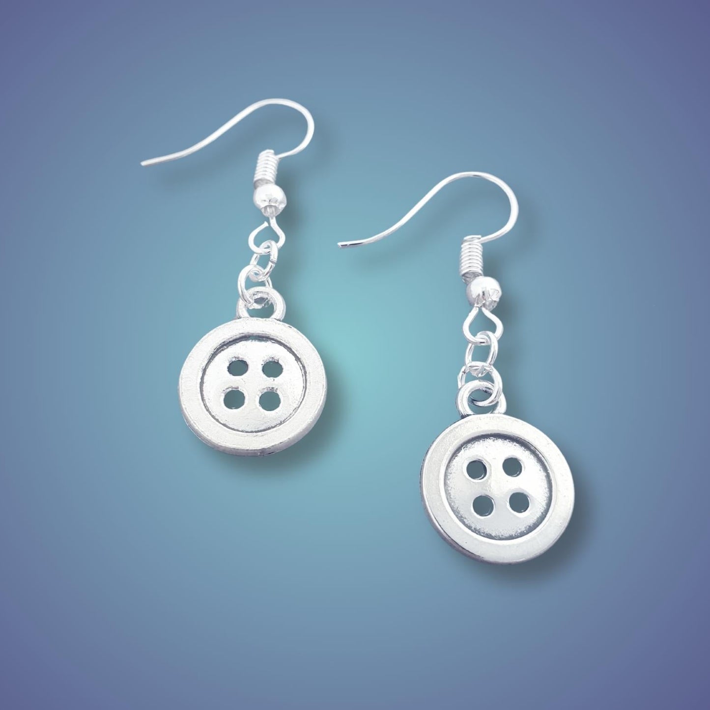 A pair of silver button dangle earrings with four holes, suspended from silver fishhook wires, shown against a blue gradient background.