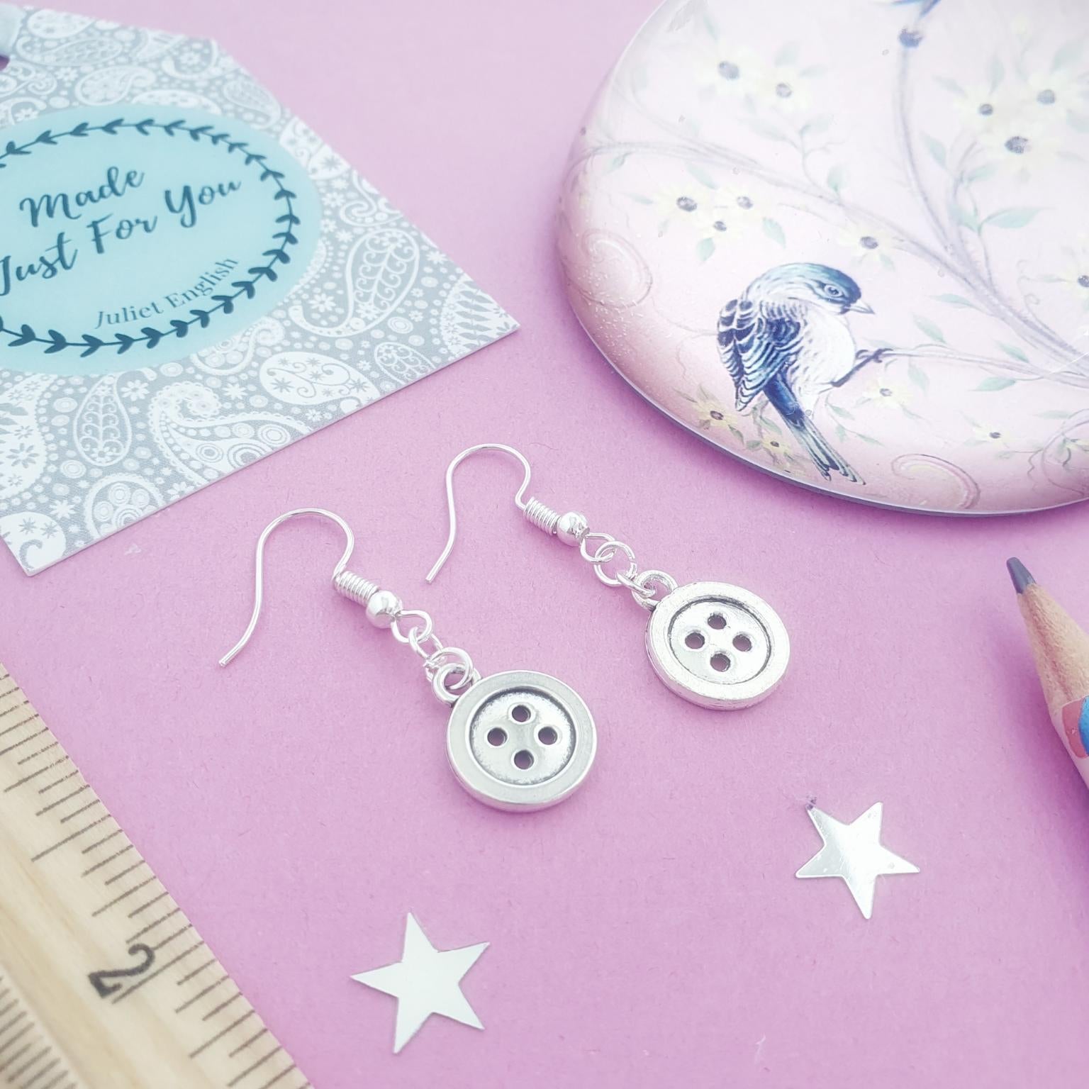 Silver button dangle earrings displayed on a pink background with a decorative gift tag, a round tin, a ruler, star cutouts, and a pencil.