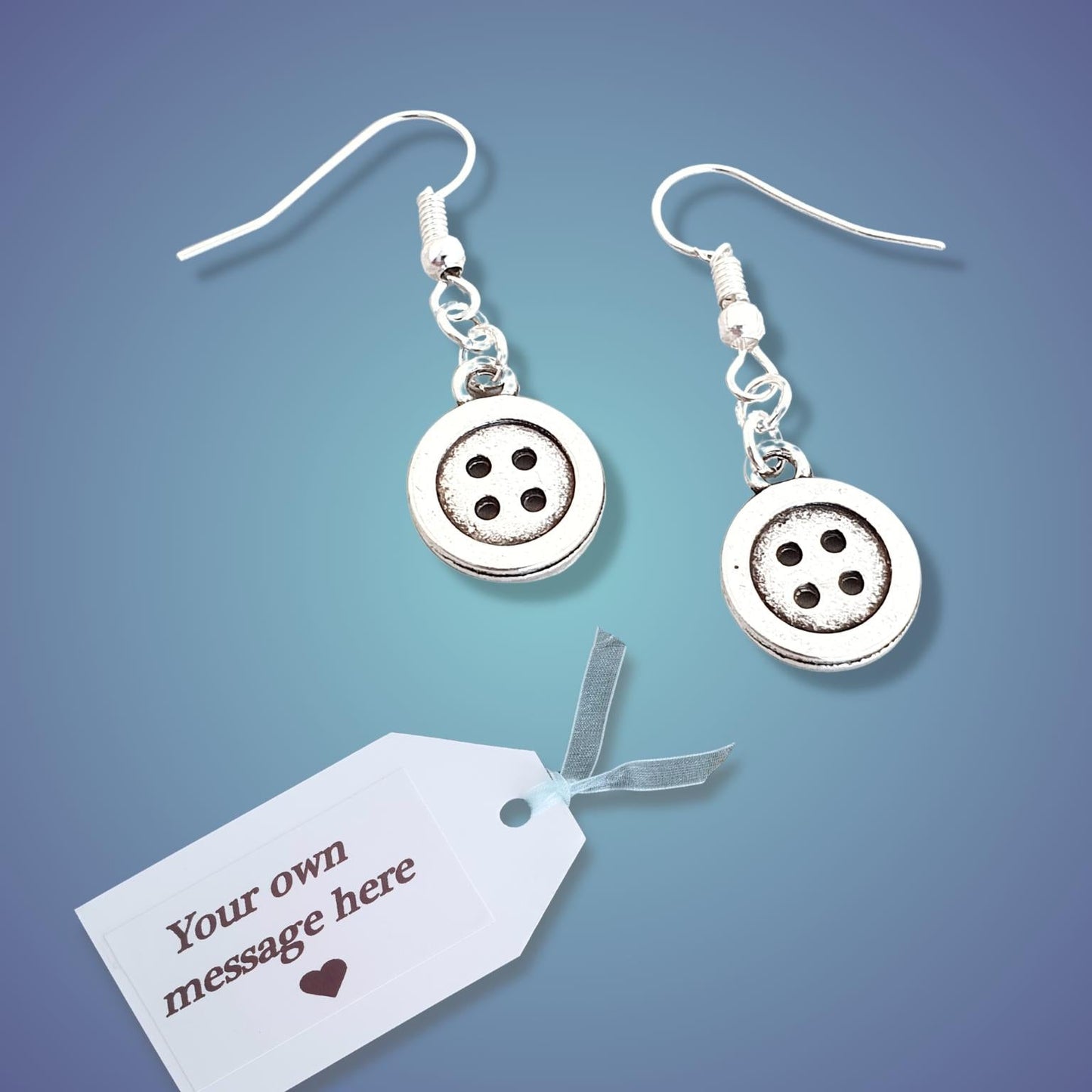 A pair of silver button dangle earrings with four-hole button charms and fishhook wires, displayed on a blue gradient background next to a white customizable gift tag.