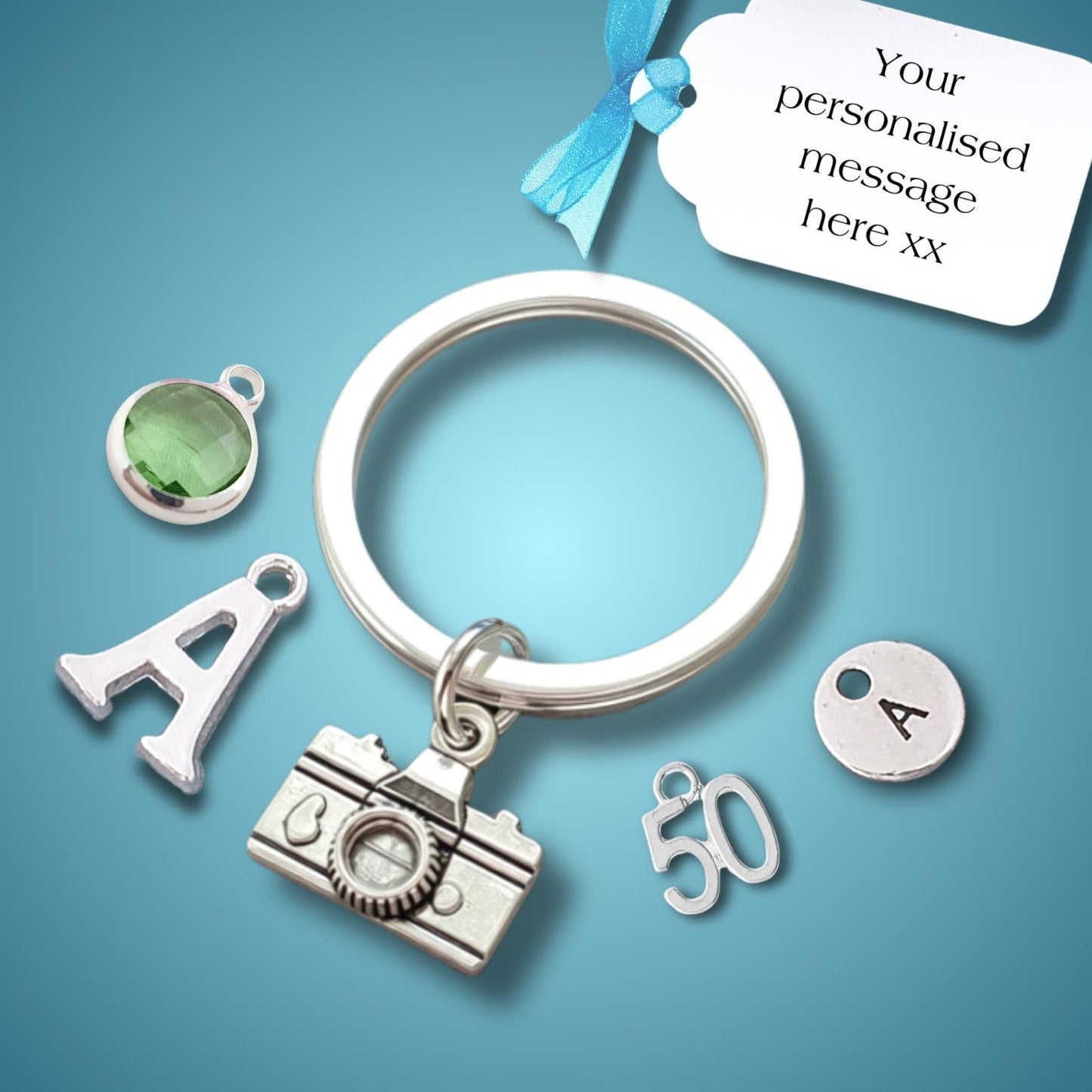 A silver keychain with a split ring, featuring multiple customizable charms including a camera, the letter 'A', a '50' charm, and a round 'A' charm. A small green oval charm is shown separately, with a white tag displaying a customizable message and a blue ribbon bow on a blue gradient background.