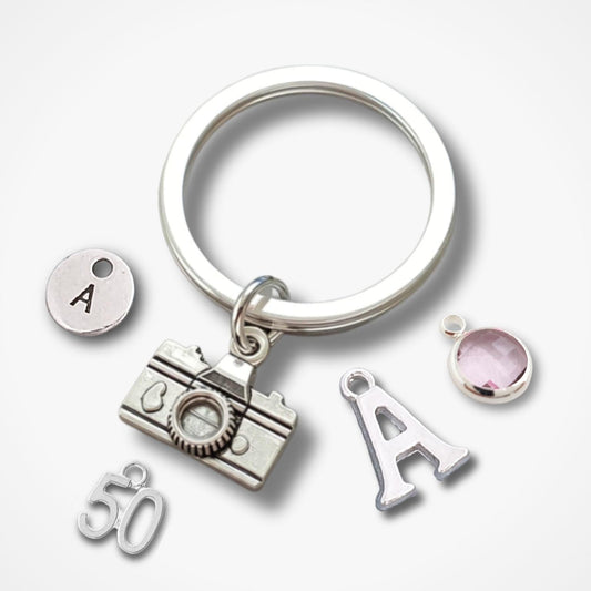 A silver keychain with a large split ring and multiple charms, including a camera, the number 50, an initial letter A charm, and a round pink gemstone charm, shown on a light gray background.