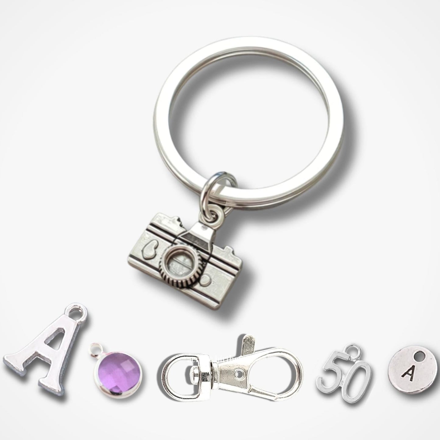 Silver camera charm keychain with a split ring, shown with five small matching jewelry components below on a light gray background.