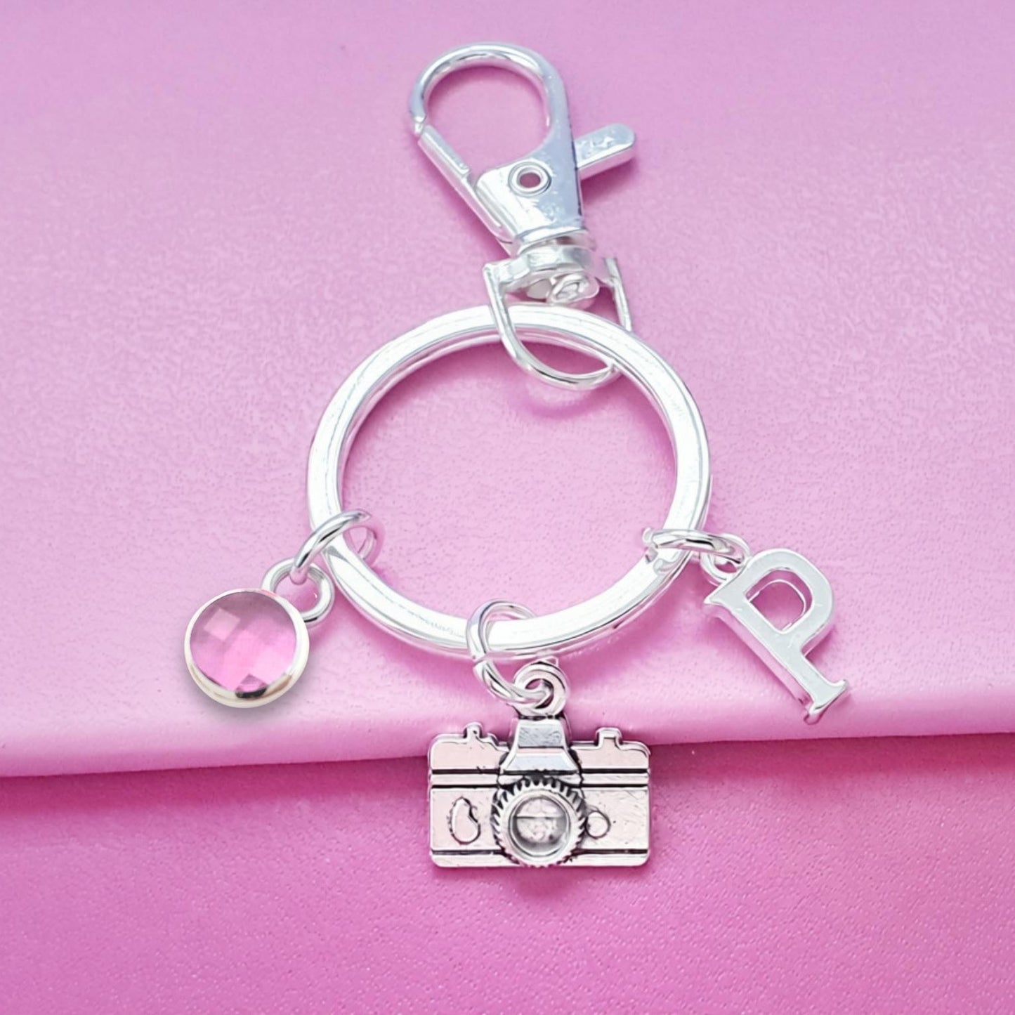 A silver-toned metal keychain featuring a lobster clasp, a split key ring, a camera charm, a letter 'P' charm, and a small round charm, displayed on a pink textured background.