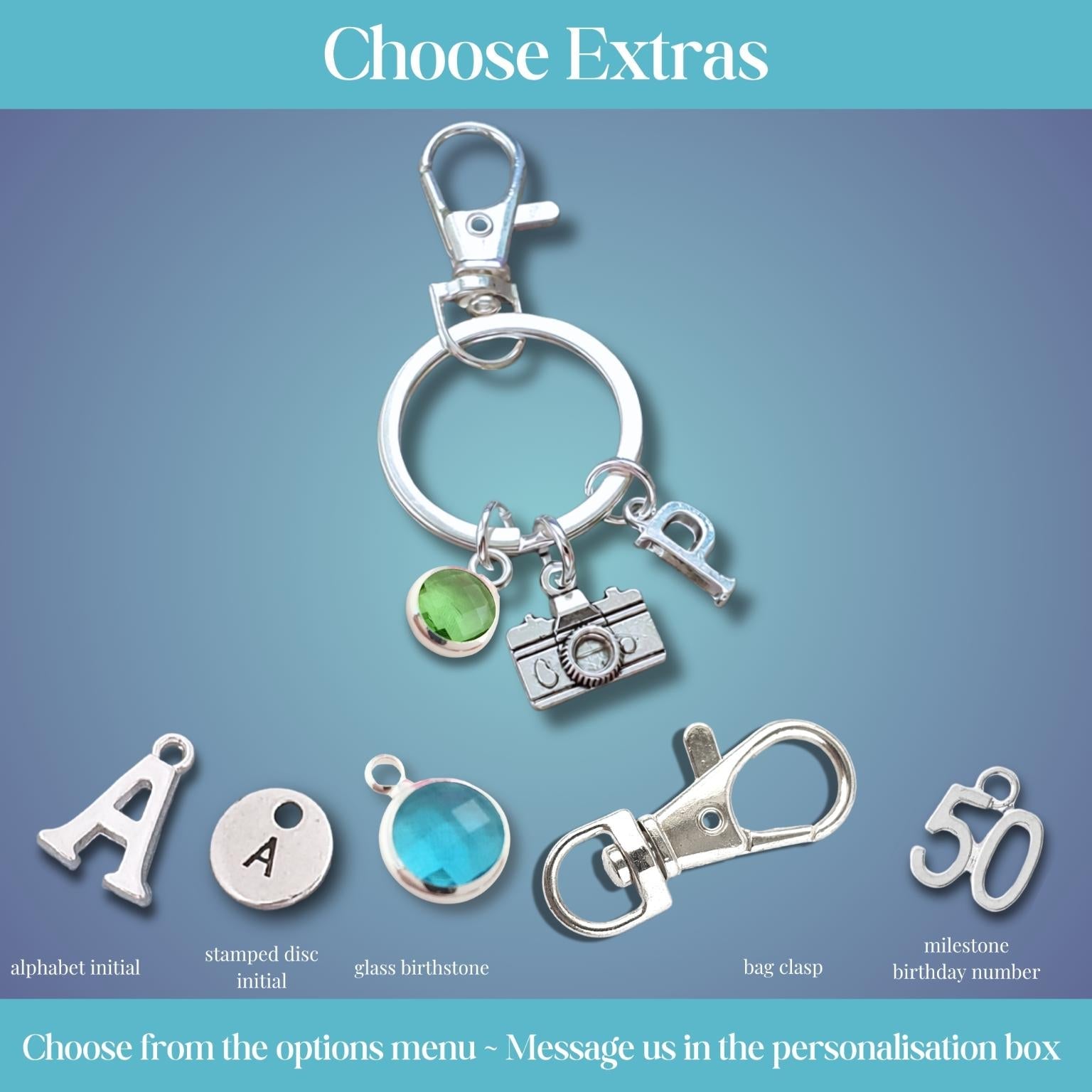 A product customization graphic showing a silver key ring with charms, including a camera, key, and green gem, alongside individual option icons for alphabet initial, stamped disc initial, glass birthstone, bag charm, and milestone birthday number on a blue gradient background with 'Choose Extras' text.