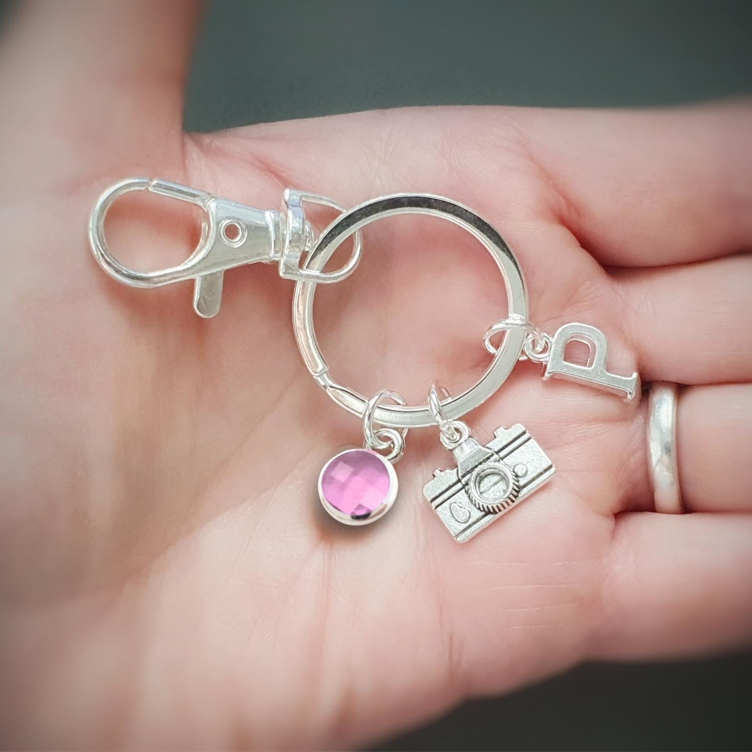 A silver keychain with a lobster clasp, split ring, a letter 'P' charm, a camera charm, and a pink round charm, held in a person's hand.