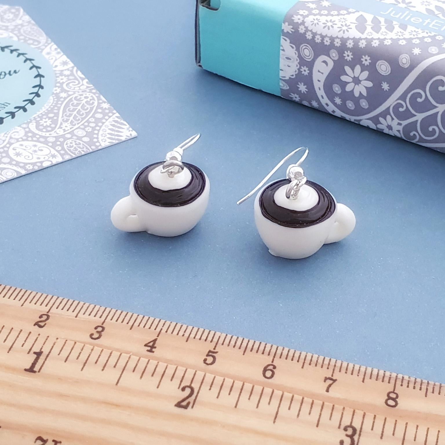 White resin coffee cup earrings with brown accents on a blue surface with a ruler for scale.