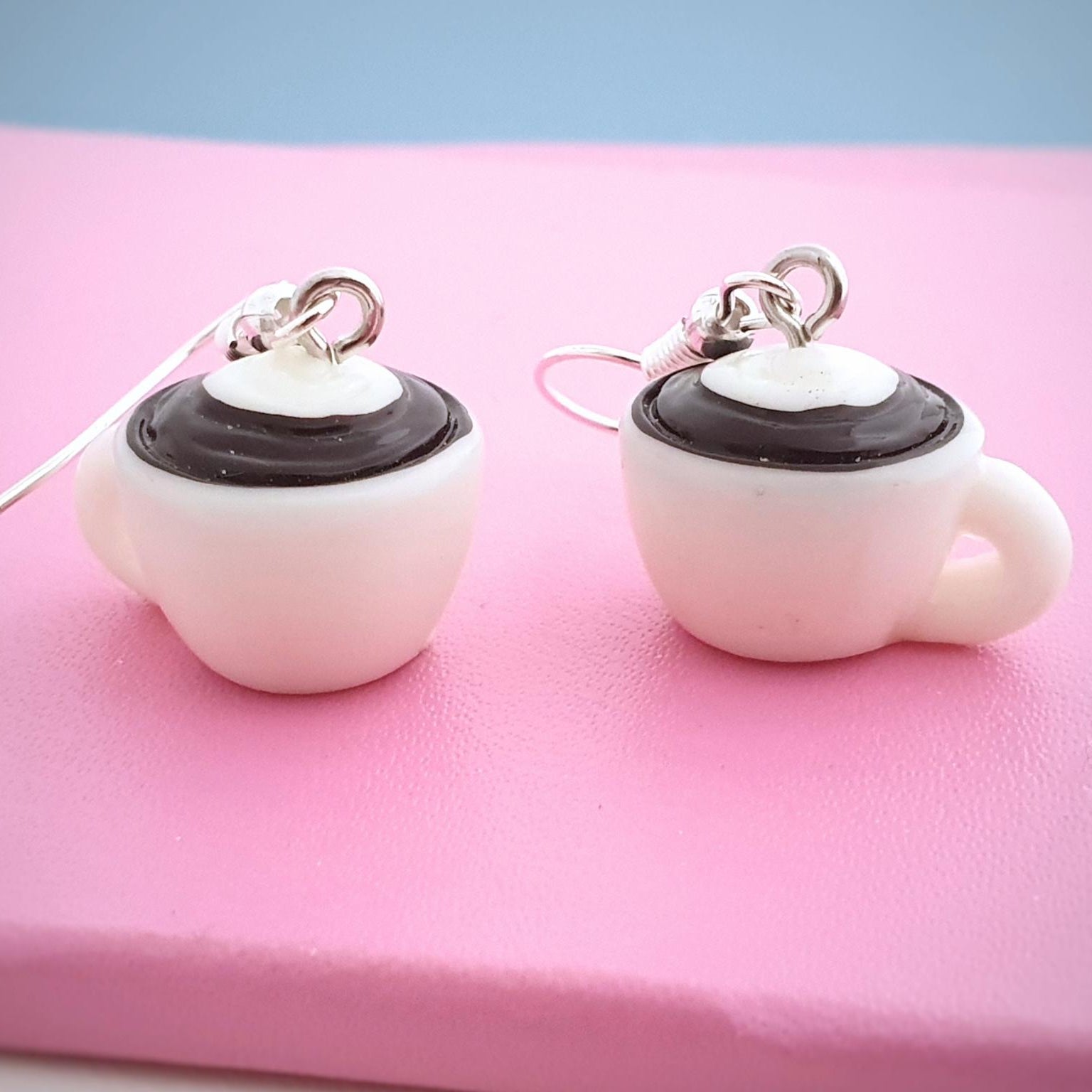 coffee-cup-shaped earrings on a pink surface with a blue background