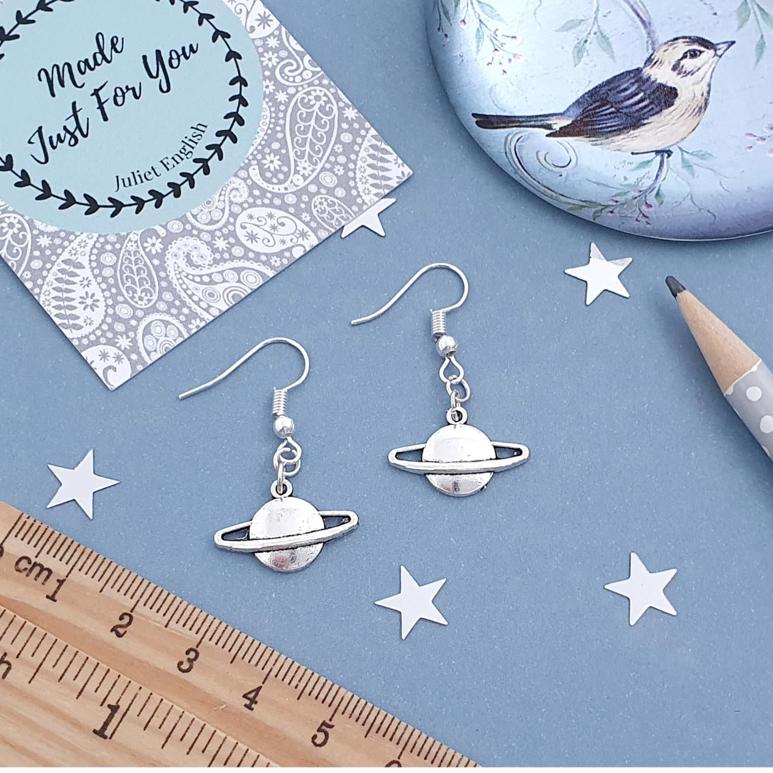 Silver planet-shaped earrings on a blue surface with a card, pencil, and ruler.