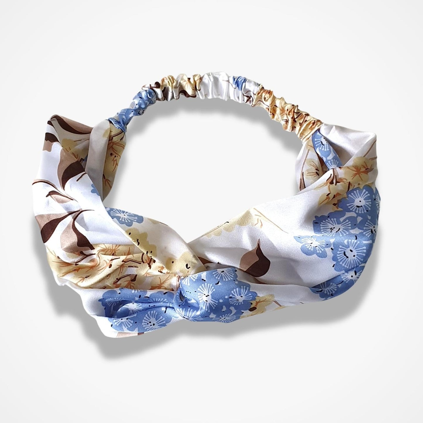 A knotted fabric headband with a white base and a pattern of blue floral motifs and brown abstract shapes, displayed on a light gray background.