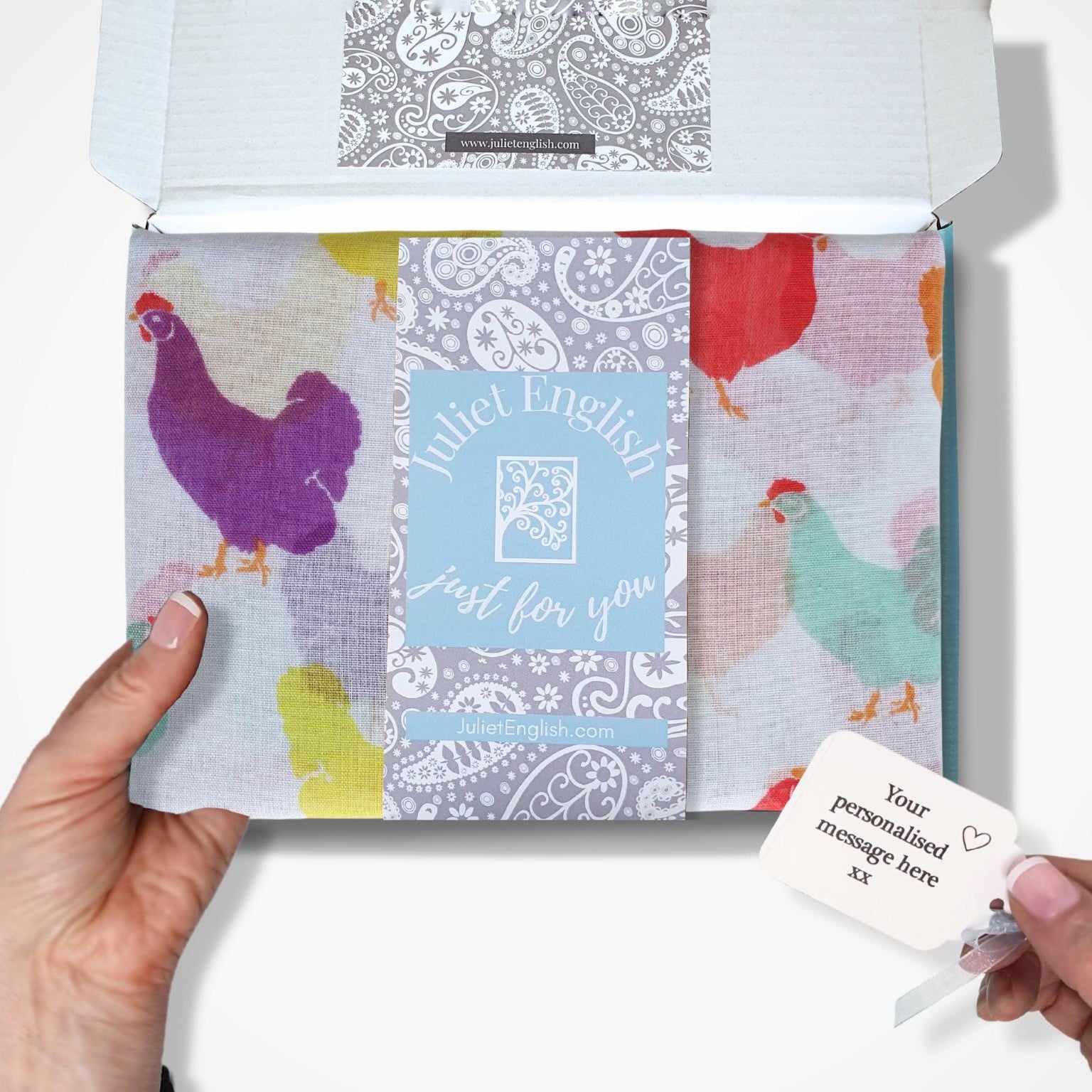 Hand holding a colourful patterned scarf in a box with a card on a white background
