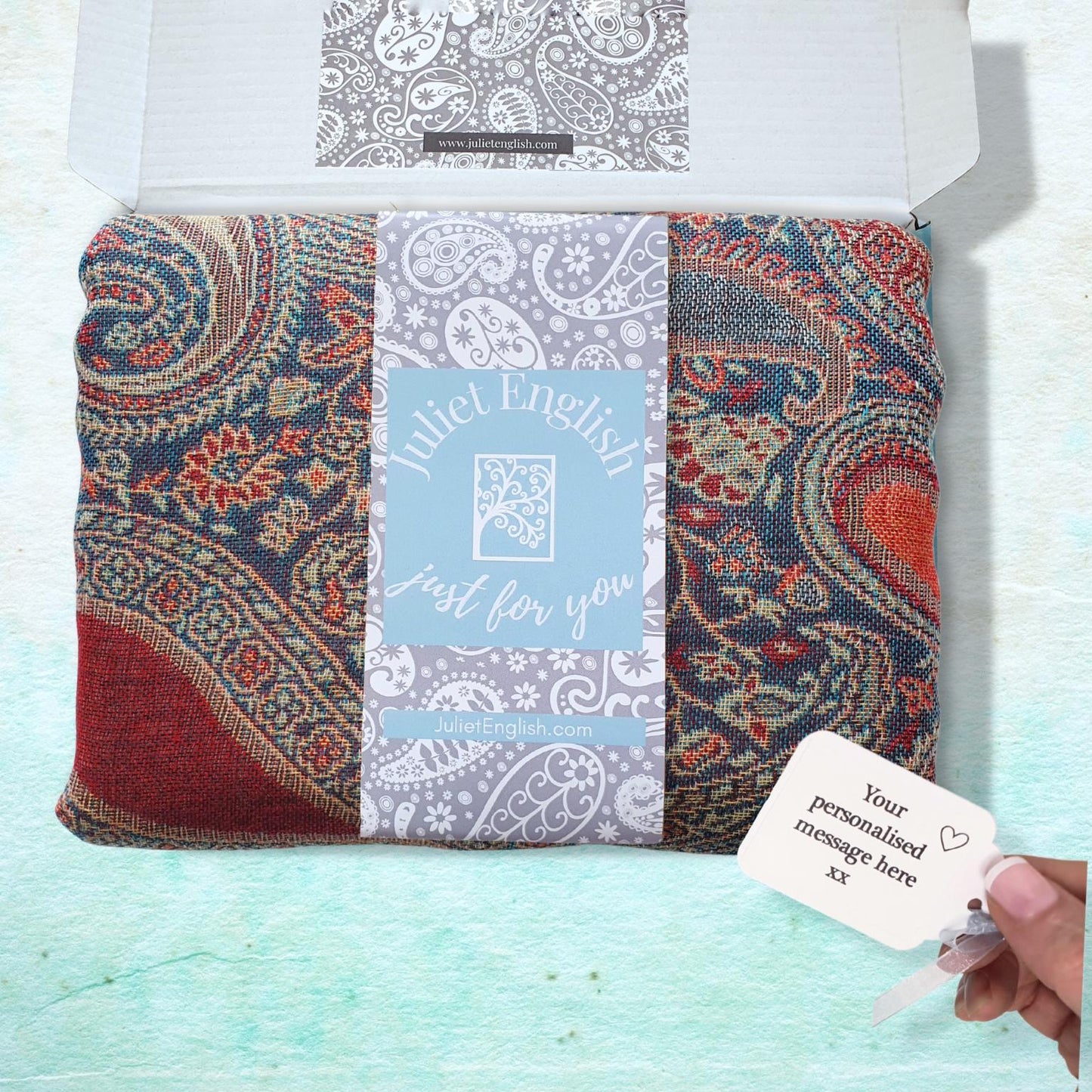 A folded paisley scarf or fabric accessory displayed in an open white gift box, with a light blue center label reading 'Juliet English just for you.' A hand is holding a small personalized tag in the lower right corner.