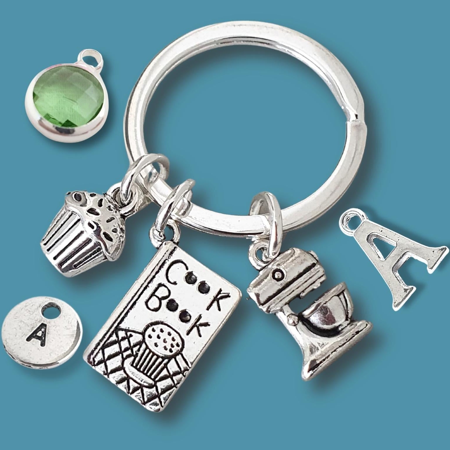 A silver plated keychain featuring multiple charms, including a green oval stone, a cupcake, a cookbook, a stand mixer, a letter 'A' charm, and a round letter 'A' disc charm, displayed on a solid teal-blue background.