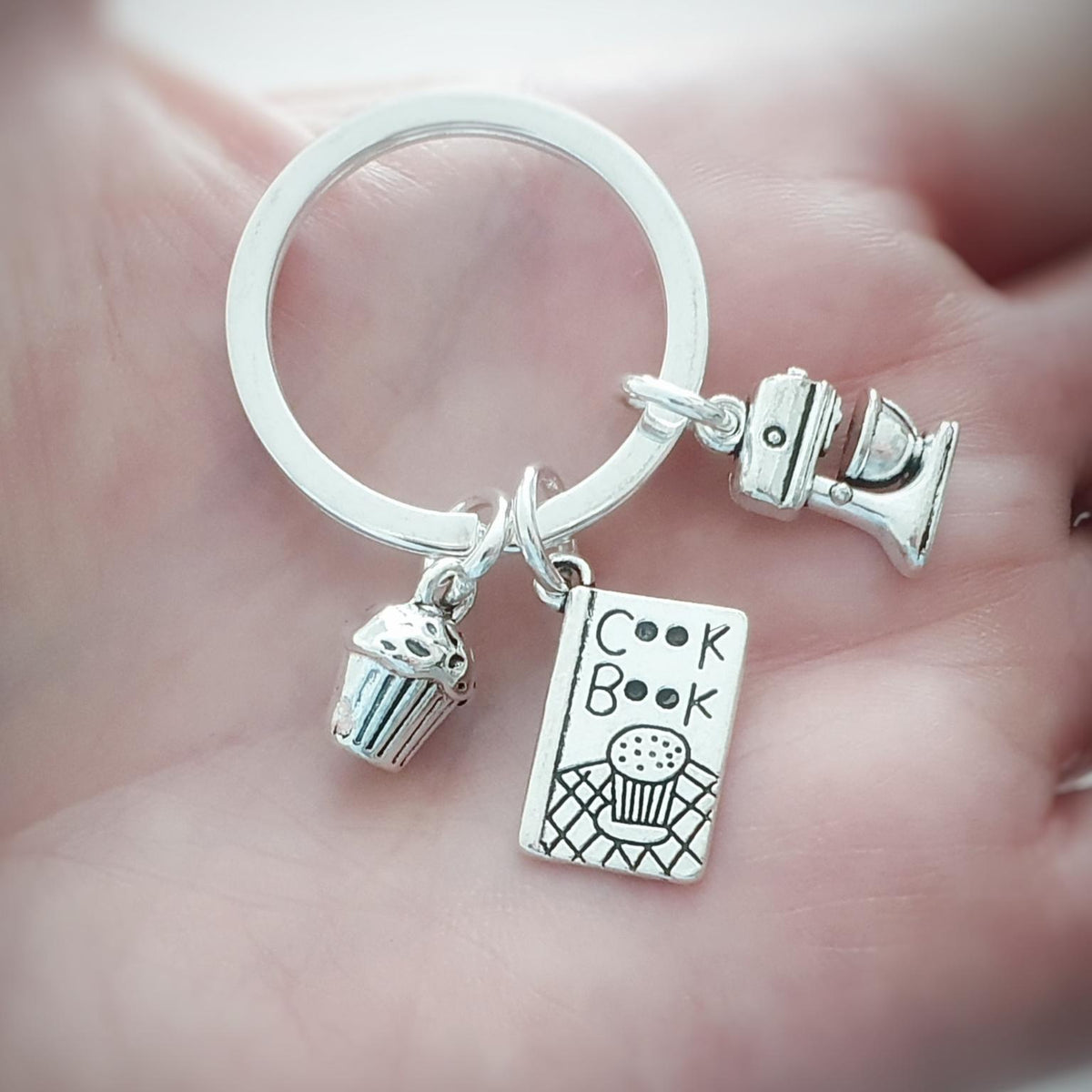 A close-up of a silver keychain held in a person's hand. The keychain has a round split ring with three charms: a cupcake charm, a book charm labeled 'Cook Book', and a small mixer charm.