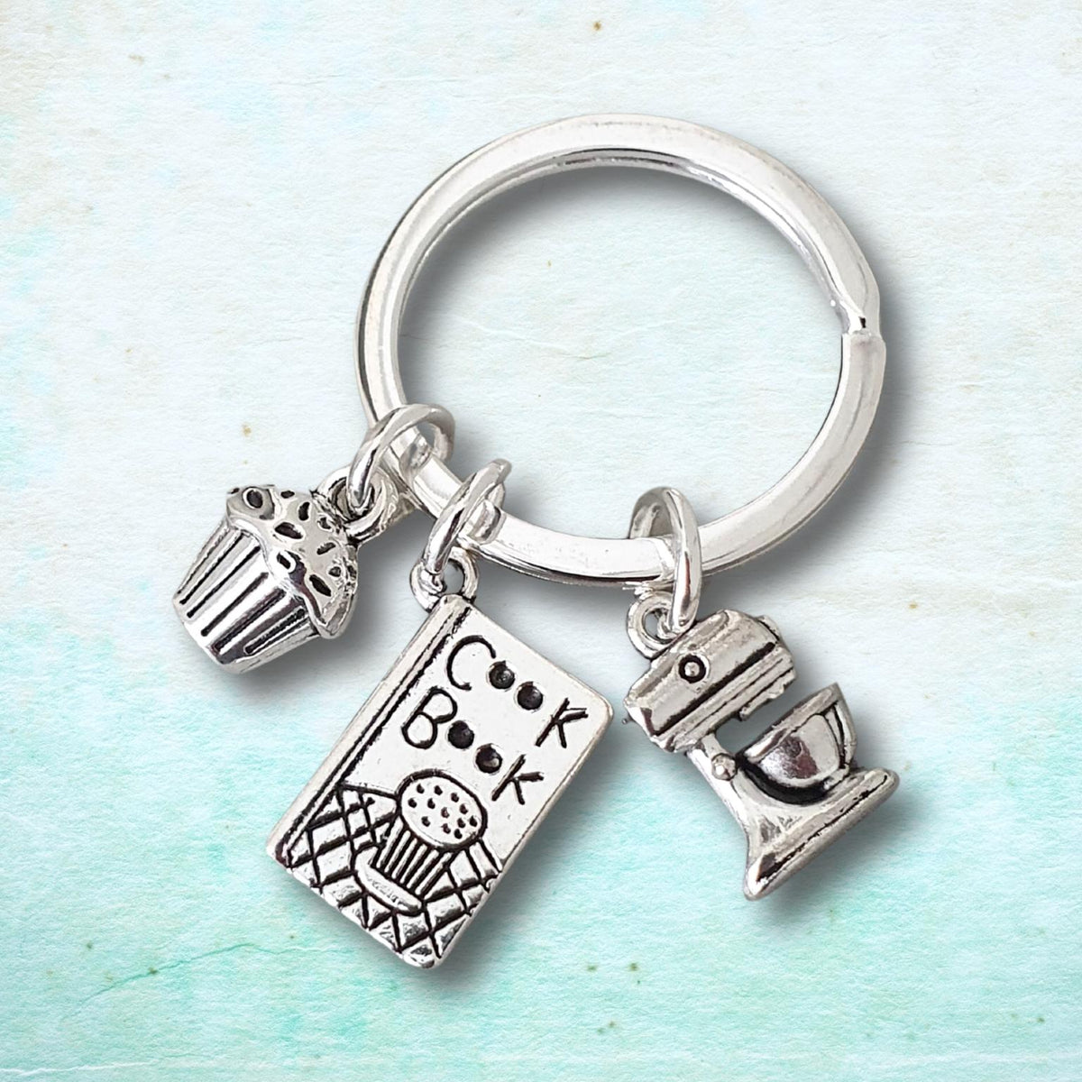 A silver-toned metal keychain featuring three baking-themed charms: a cupcake, a cookbook with the word 'Cook Book' engraved, and a stand mixer, shown on a light blue textured background.
