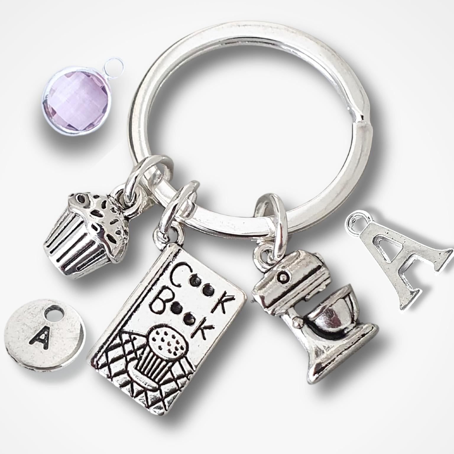A silver keychain with multiple charms, including a cupcake, a book labeled 'cook book', a mixer and letter 'A' charms, alongside a small lavender faceted gemstone, shown on a light gray background.