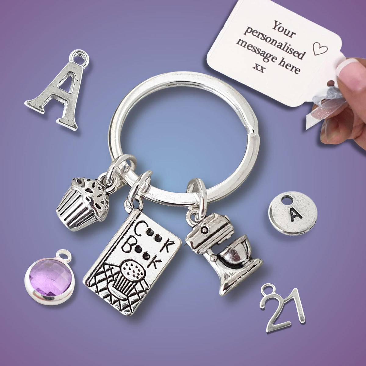 A silver keychain with multiple charms, including a cupcake, a book, a mixer, a letter 'A', a round 'A' charm, and a purple oval gem, shown on a purple gradient background. A hand is holding a white tag that reads 'Your personalised message here xx'.
