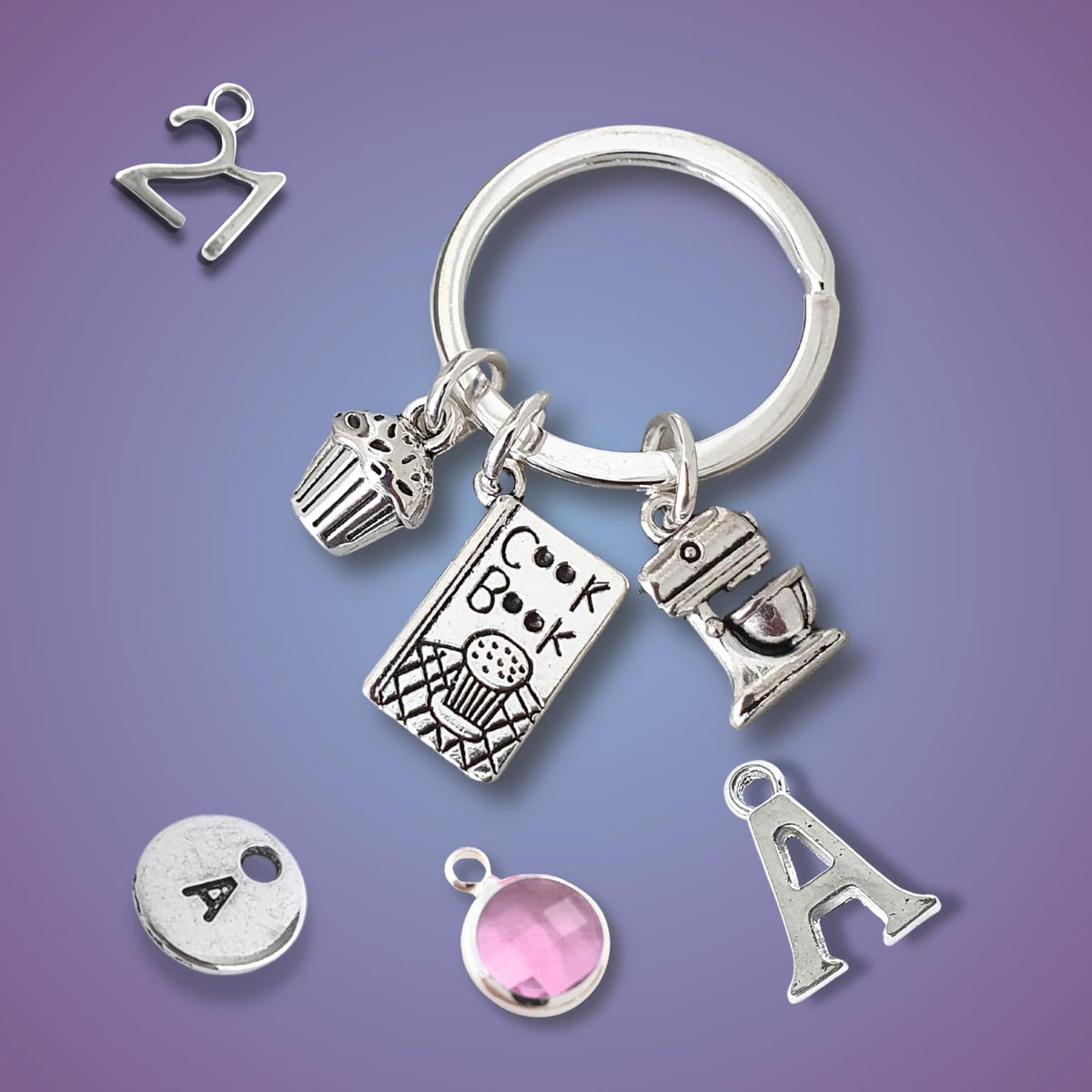 A silver keychain with multiple charms, including a cupcake, a cookbook, a mixerand an initial 'A' charm, and a pink gemstone charm, displayed on a purple gradient background.