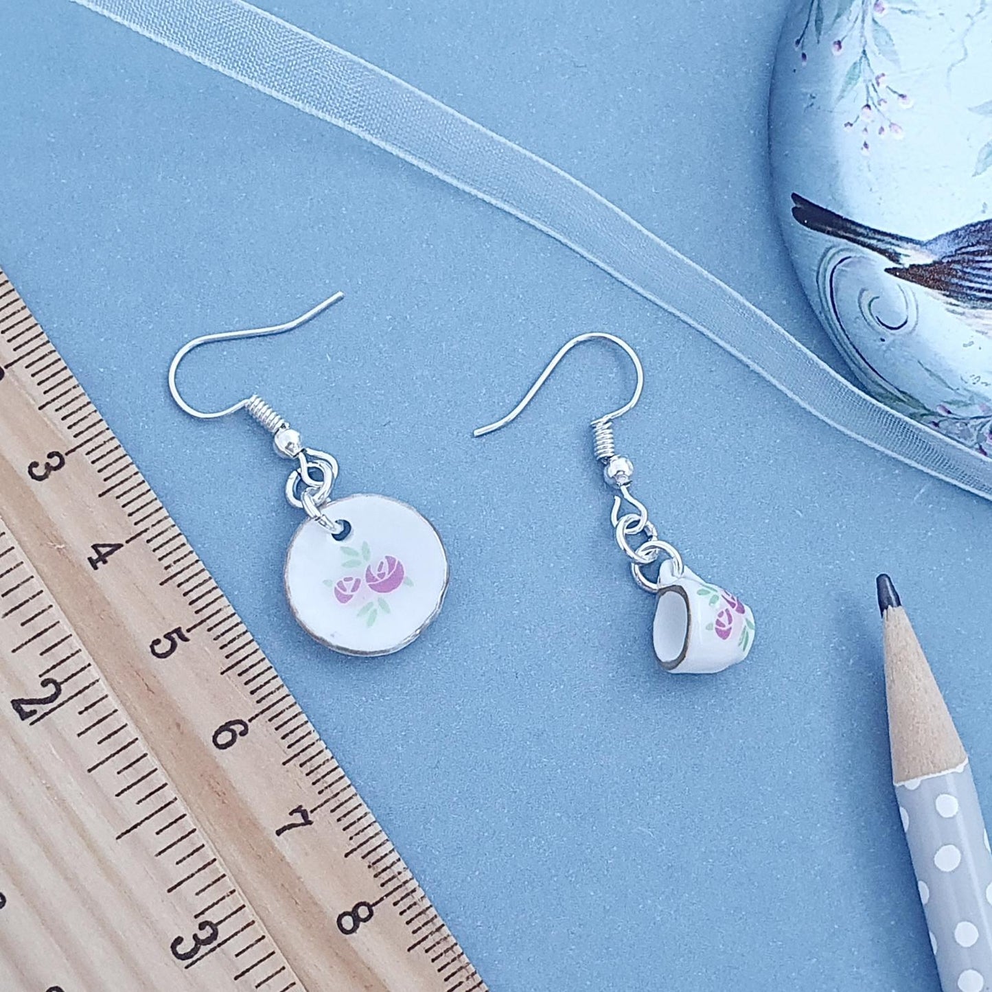 Pair of cup and saucer earrings with floral designs on a blue surface with a ruler and pencil for scale.