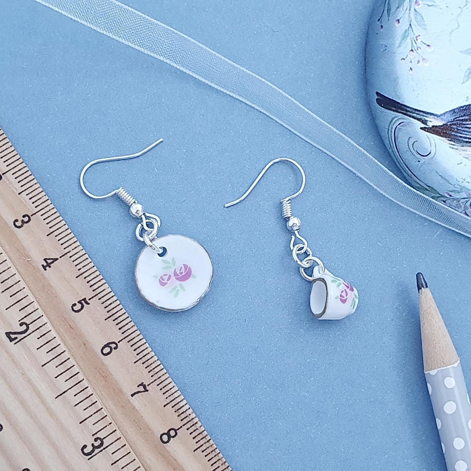 Pair of cup and saucer earrings with floral designs on a blue surface with a ruler and pencil for scale.